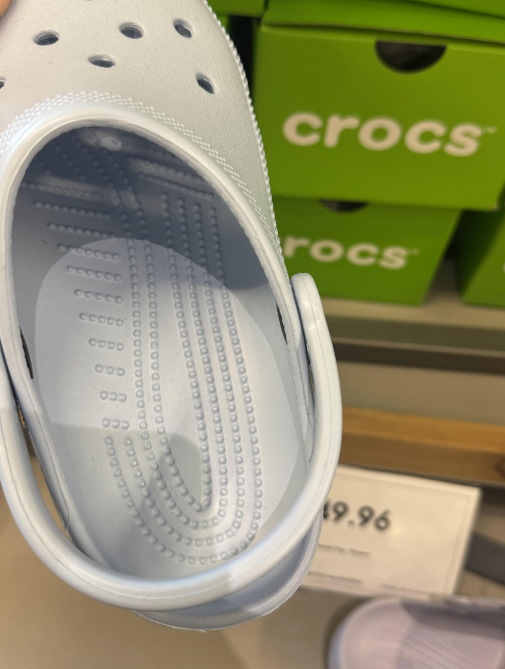 A close-up of a Croc sandal held above a store display shows the detailed insole texture and stacked boxes labeled "crocs" in the background