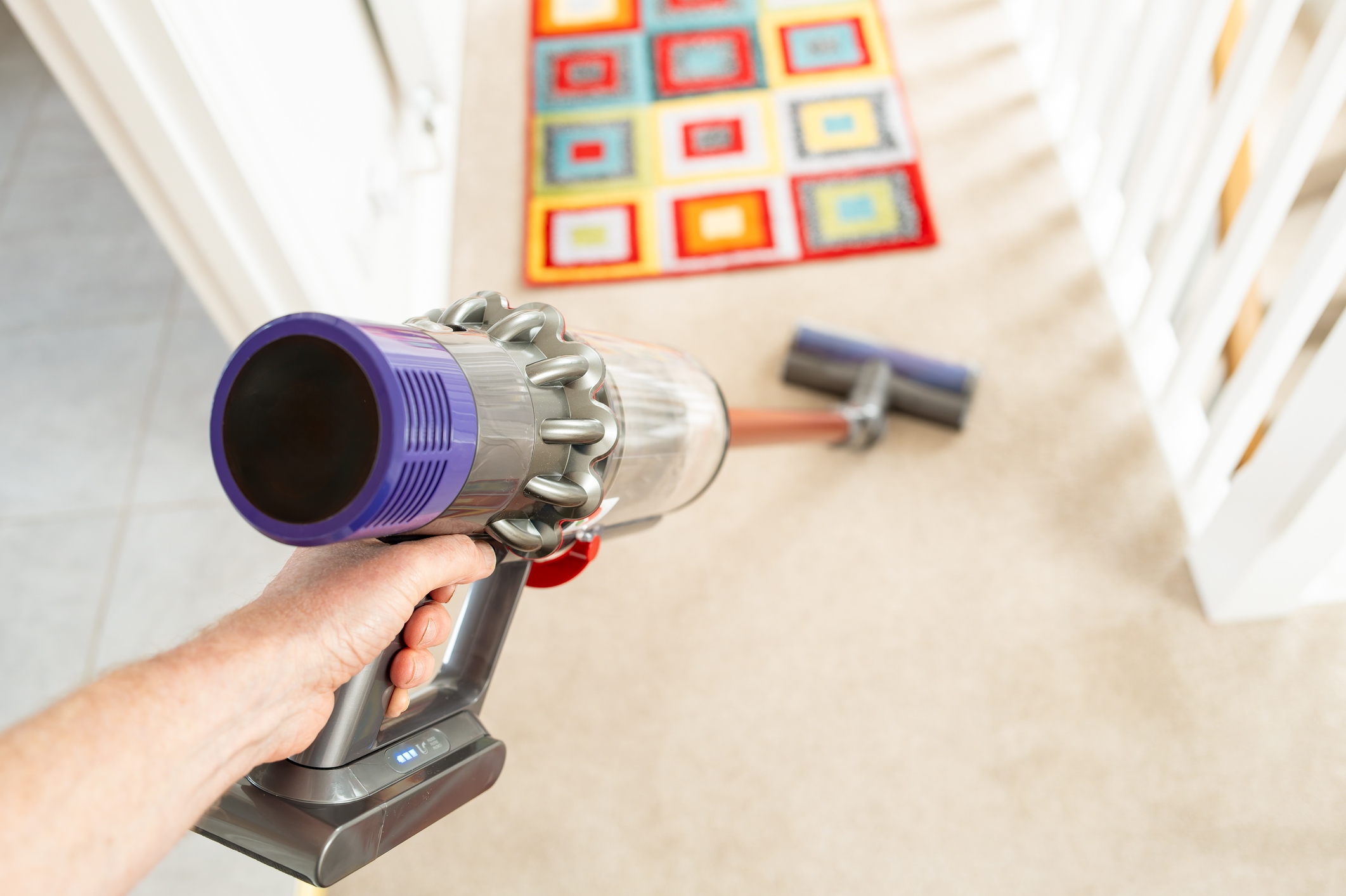 Hand holding a cordless vacuum, cleaning a carpet with a colorful geometric pattern rug nearby