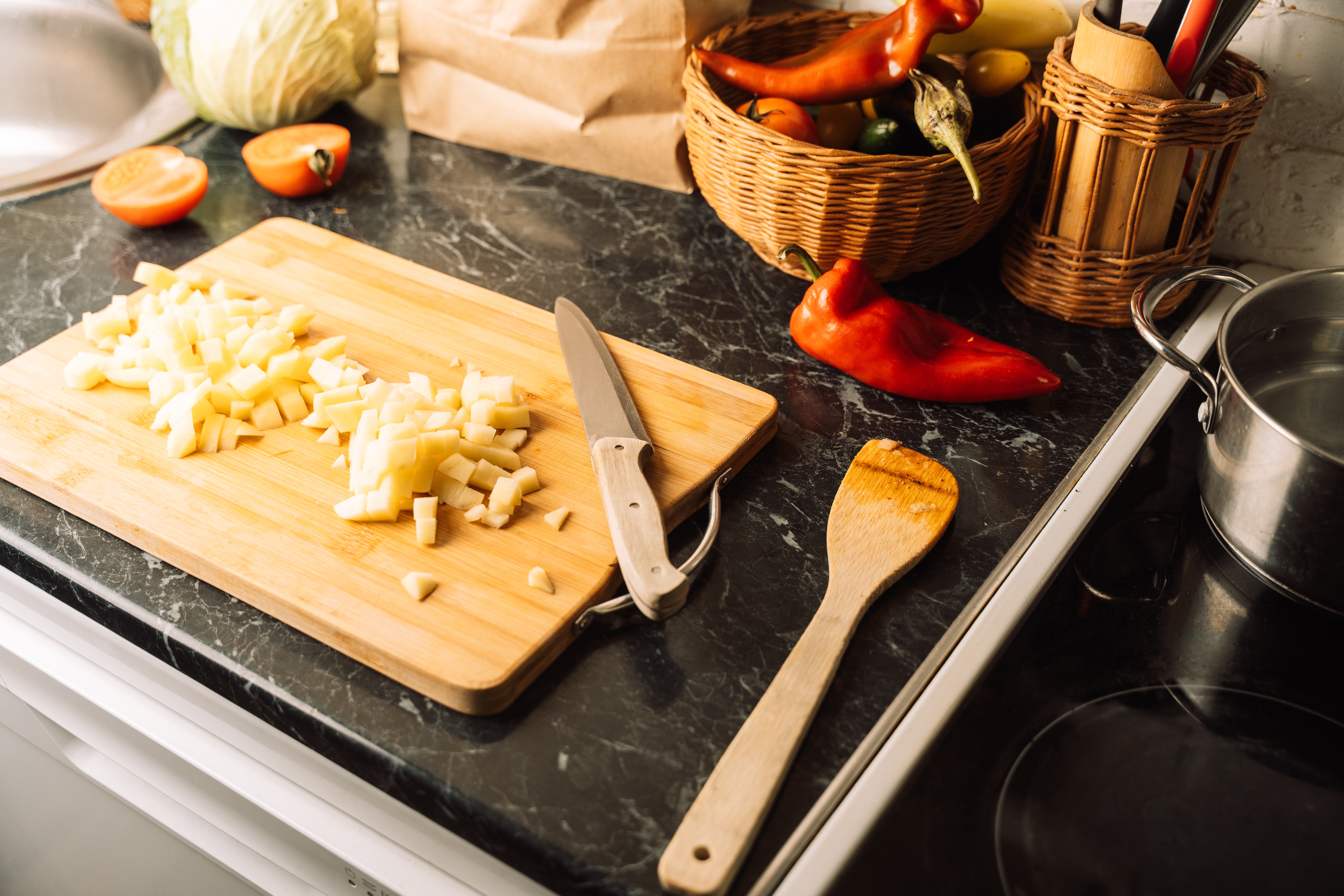 Skjær grønnsaker på et trebrett med en kniv, omgitt av en kurv med råvarer, en treskje og en gryte på en kjøkkenbenk