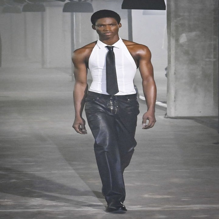 Model on a runway wearing a sleeveless white top with a black tie and leather pants