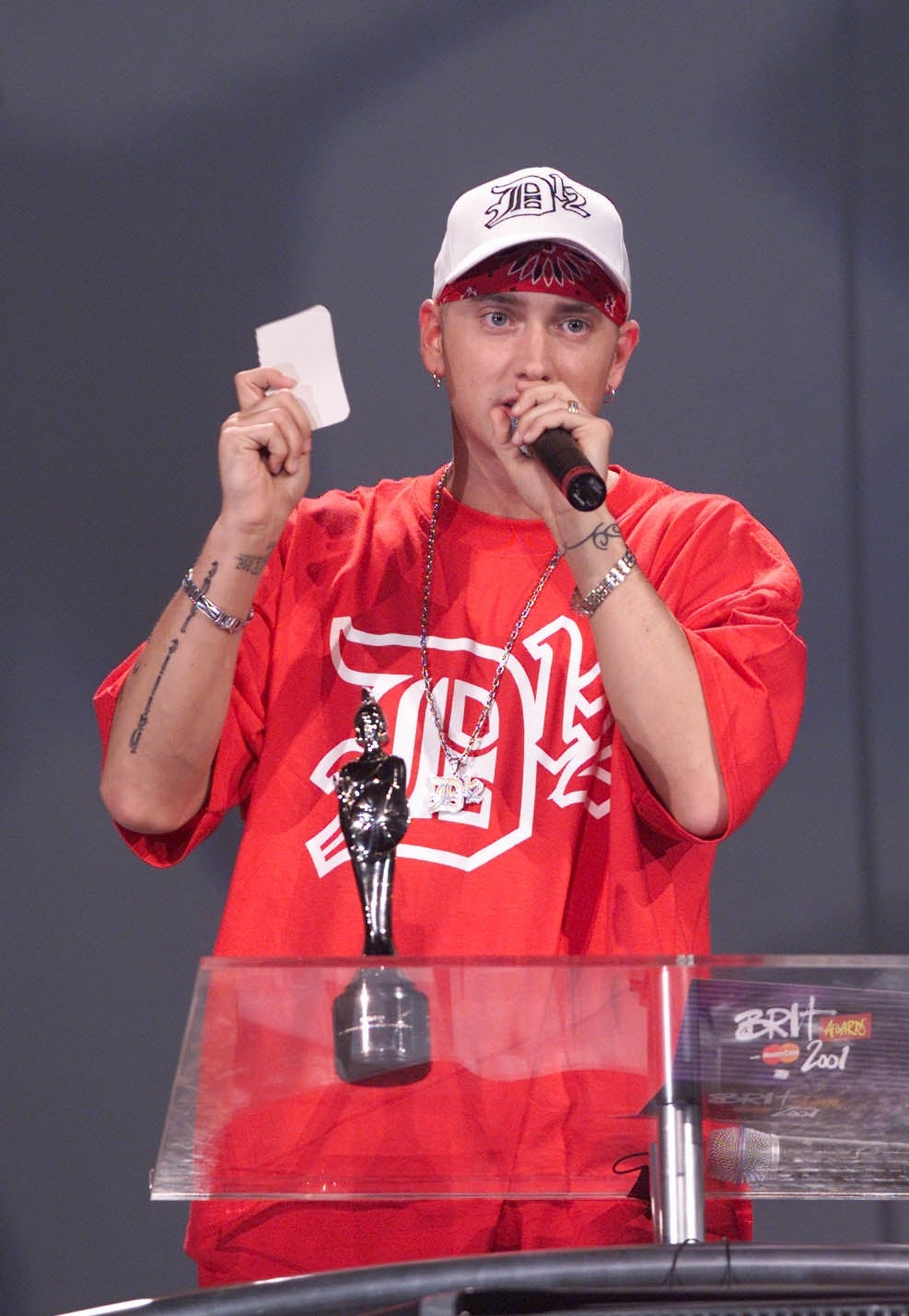 14 Celeb Feuds That Were SO One-Sided, They Were Basically Beefing With A Brick Wall 4 Celebrity on stage holding a card and trophy, wearing a casual red shirt and cap at an award ceremony