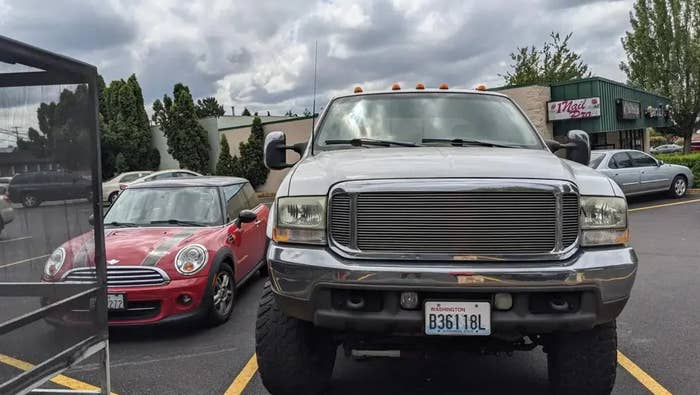 A small car is parked next to a large pickup truck in a parking lot, showing a size comparison. The image is on a Reddit page about cars