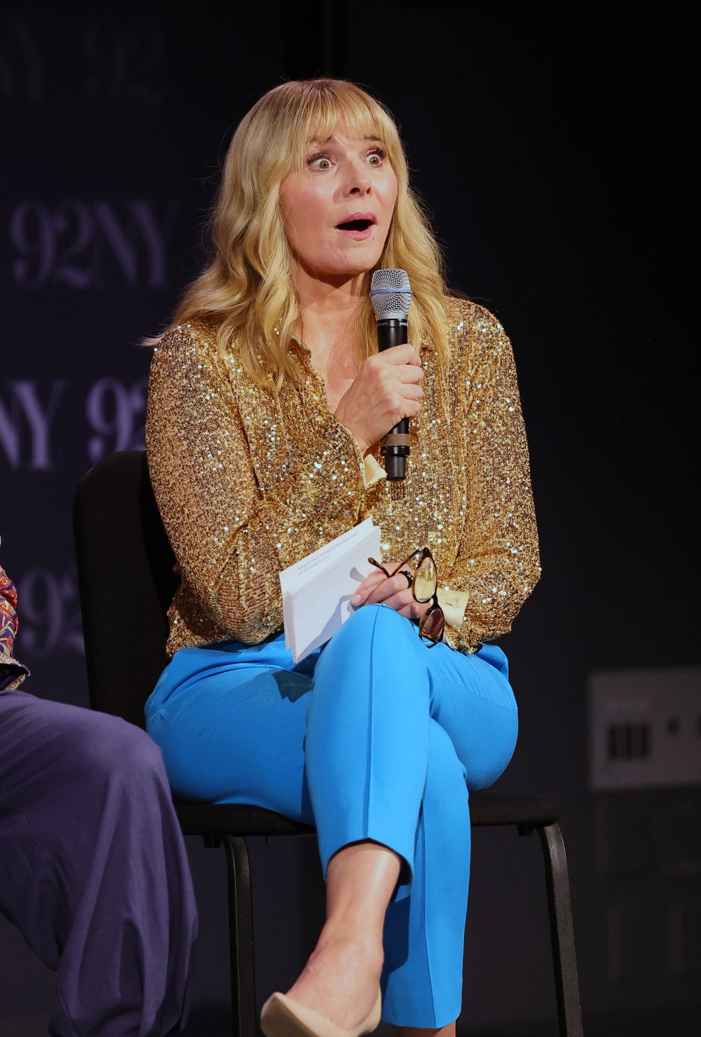 14 Celeb Feuds That Were SO One-Sided, They Were Basically Beefing With A Brick Wall 36 A person on stage holds a microphone and paper, wearing a sparkling top and blue pants, appearing to speak or engage with an audience