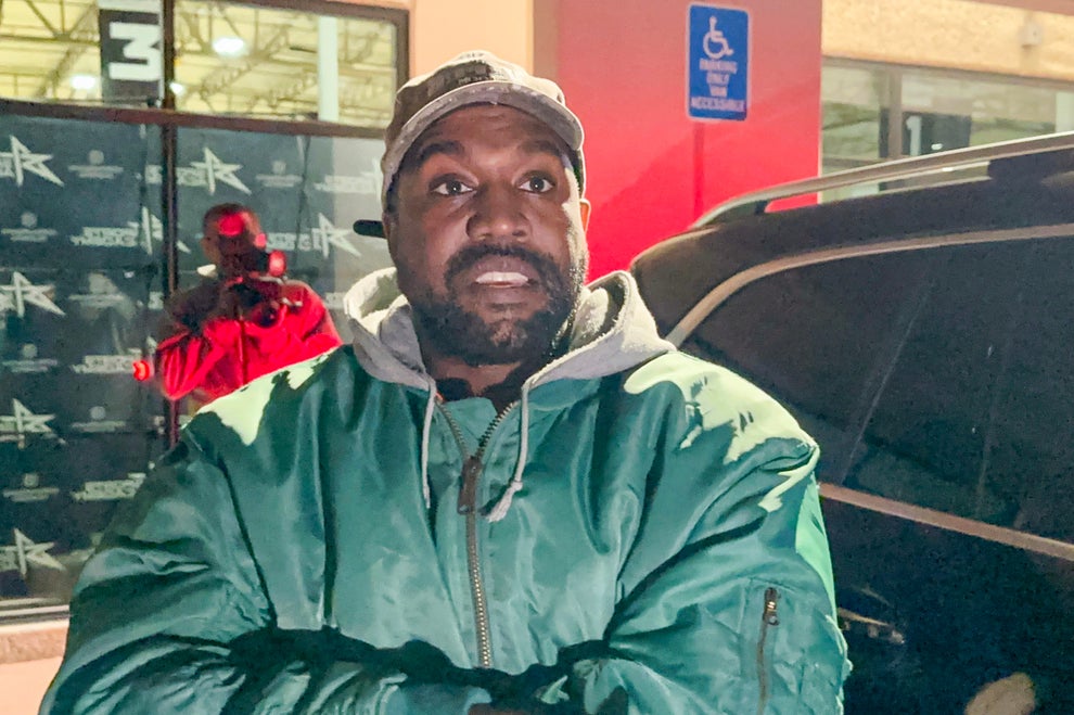 14 Celeb Feuds That Were SO One-Sided, They Were Basically Beefing With A Brick Wall 20 A person in a green jacket and cap stands in a parking lot near a vehicle. A figure in a red outfit is behind them, next to a building