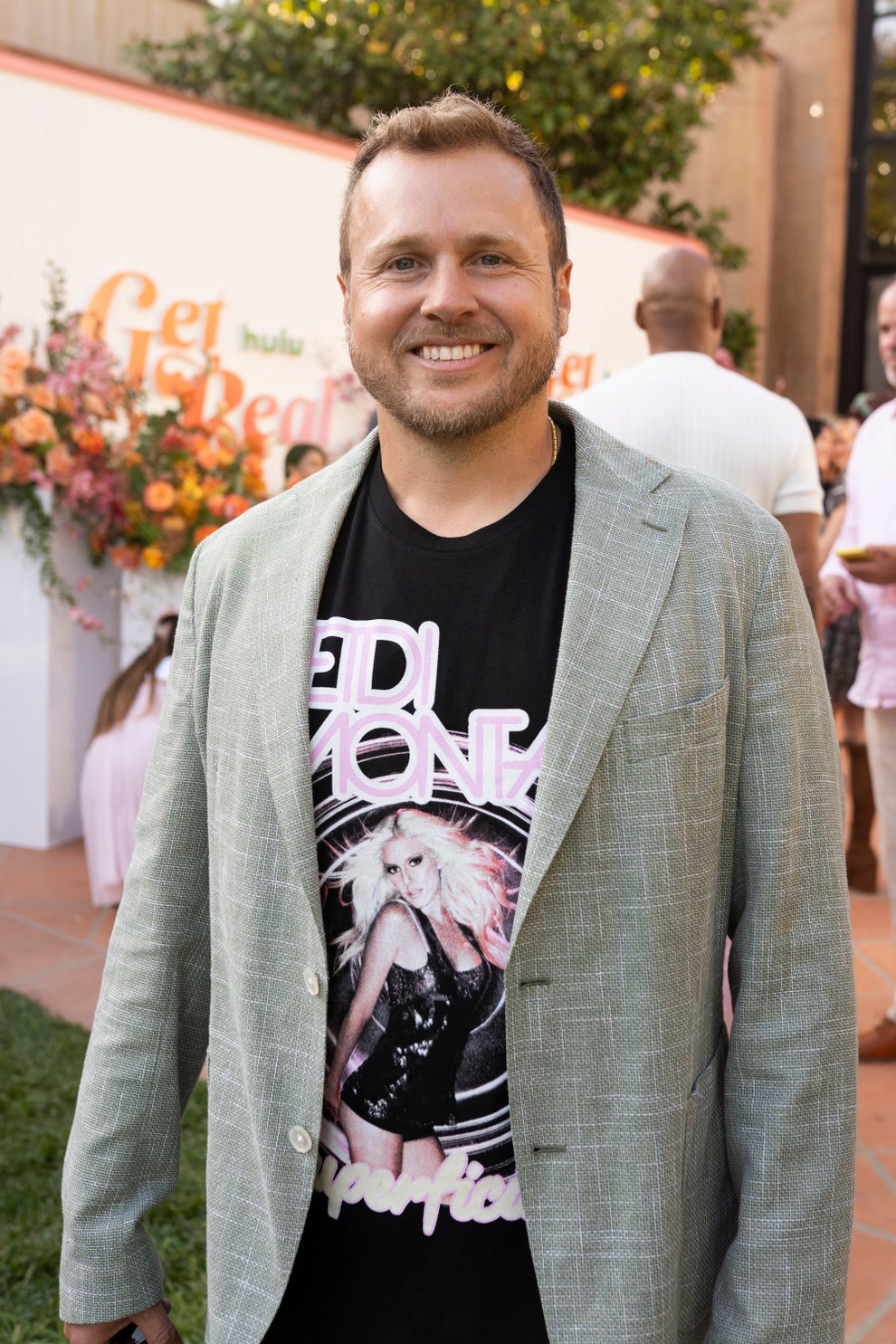 14 Celeb Feuds That Were SO One-Sided, They Were Basically Beefing With A Brick Wall 44 Man in a graphic T-shirt and casual blazer posing outdoors, smiling at an event with floral decor in the background