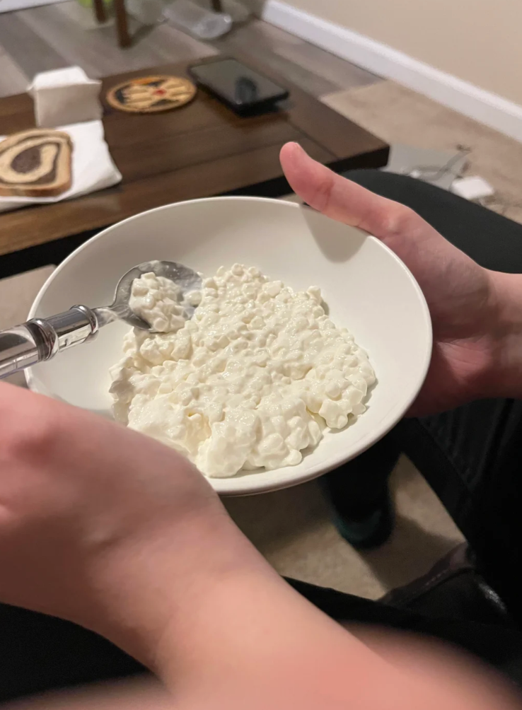 Person holding a bowl of cottage cheese in front of a computer showing a Reddit post discussing eating cottage cheese straight from a bowl