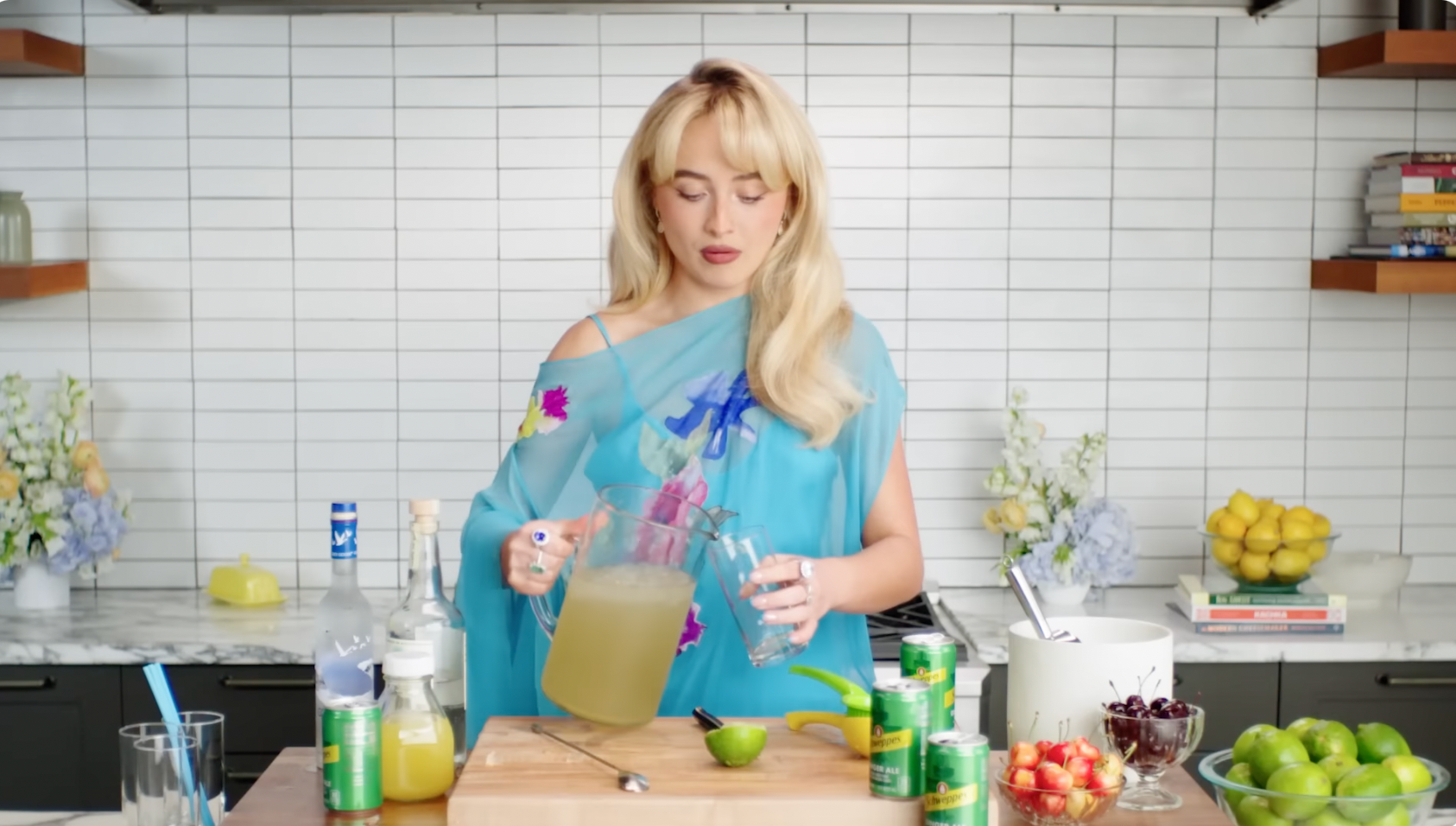 Person in a stylish off-shoulder dress pours a drink in a modern kitchen, surrounded by various ingredients and fresh fruit