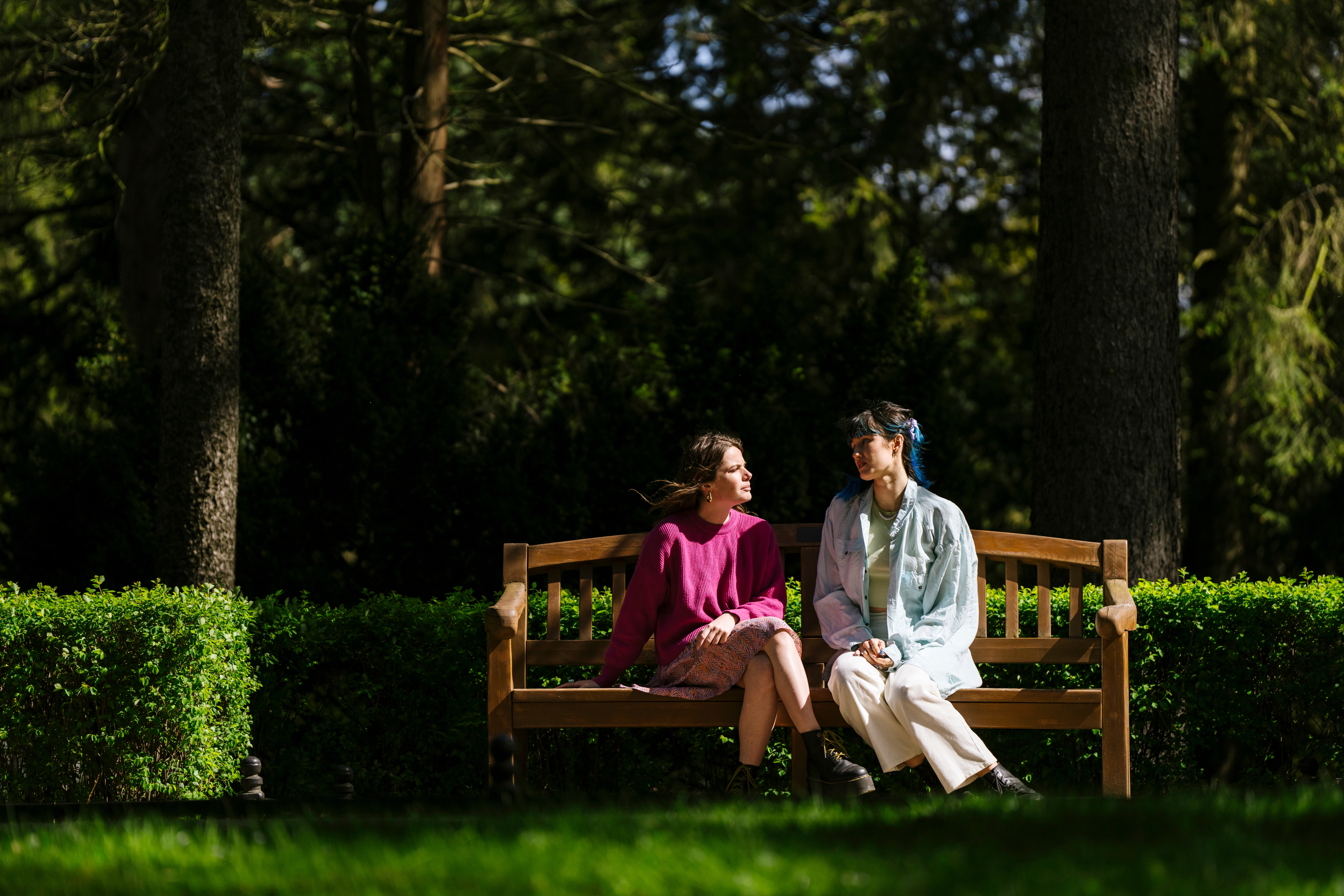 To personer sitter på en trebenk i en park, engasjert i en vennlig samtale, omgitt av frodige grøntområder og høye trær