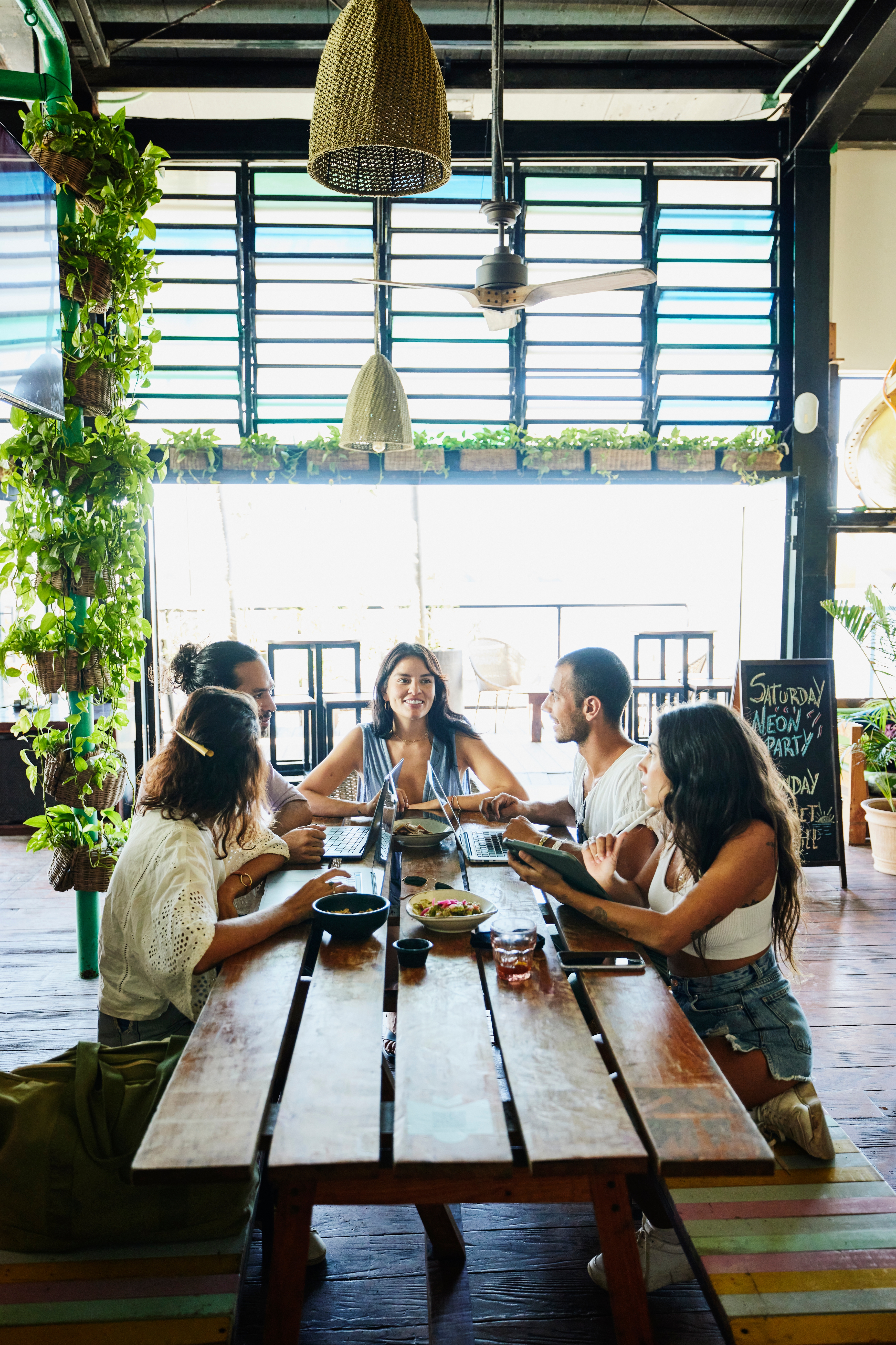 Venner nyter et måltid rundt et trebord i en lys, plantefylt kafé, og skaper en avslappet og vennlig atmosfære