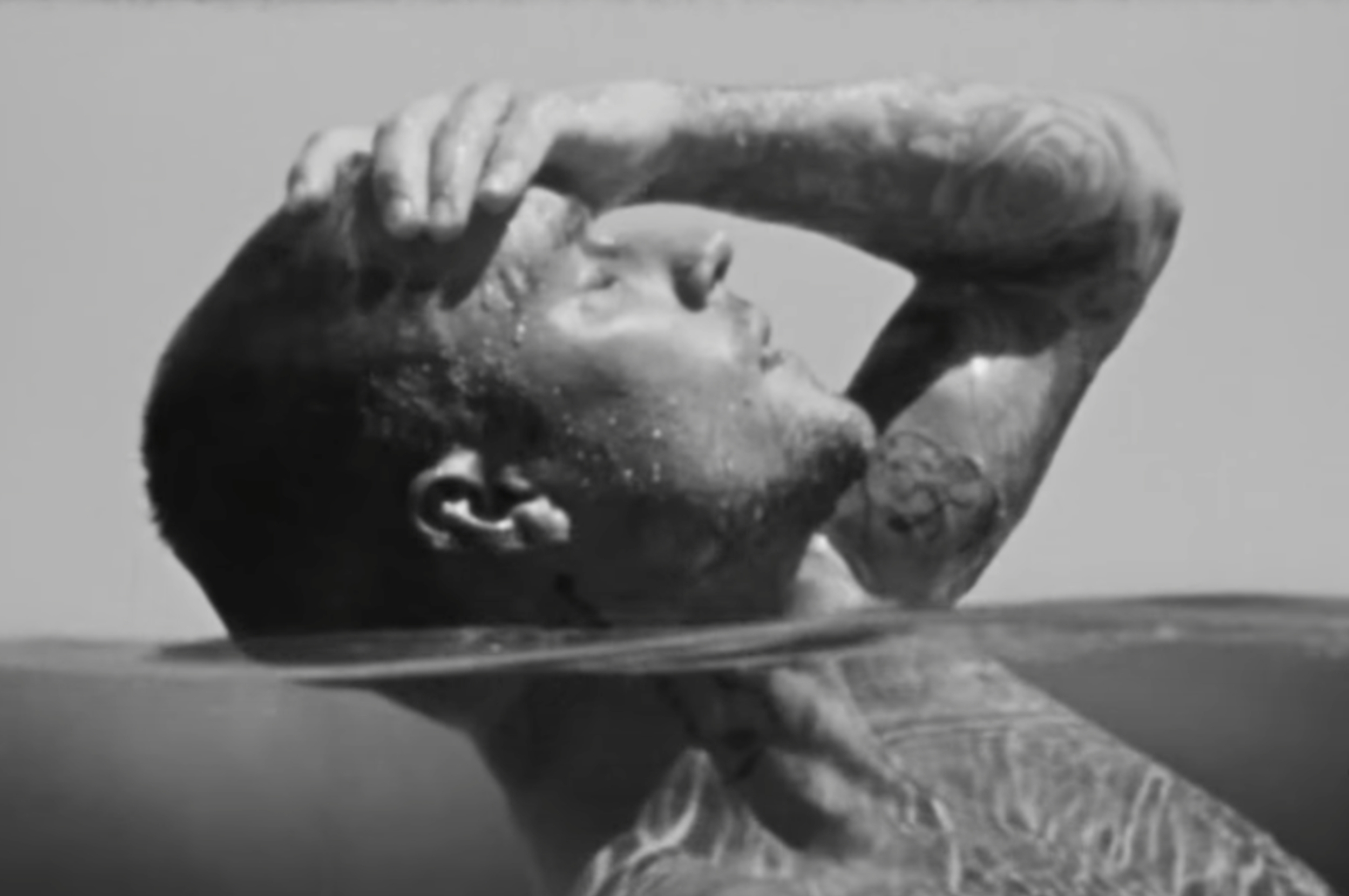 Person partially submerged in water, with tattoos visible, resting their hand on their forehead, appearing relaxed