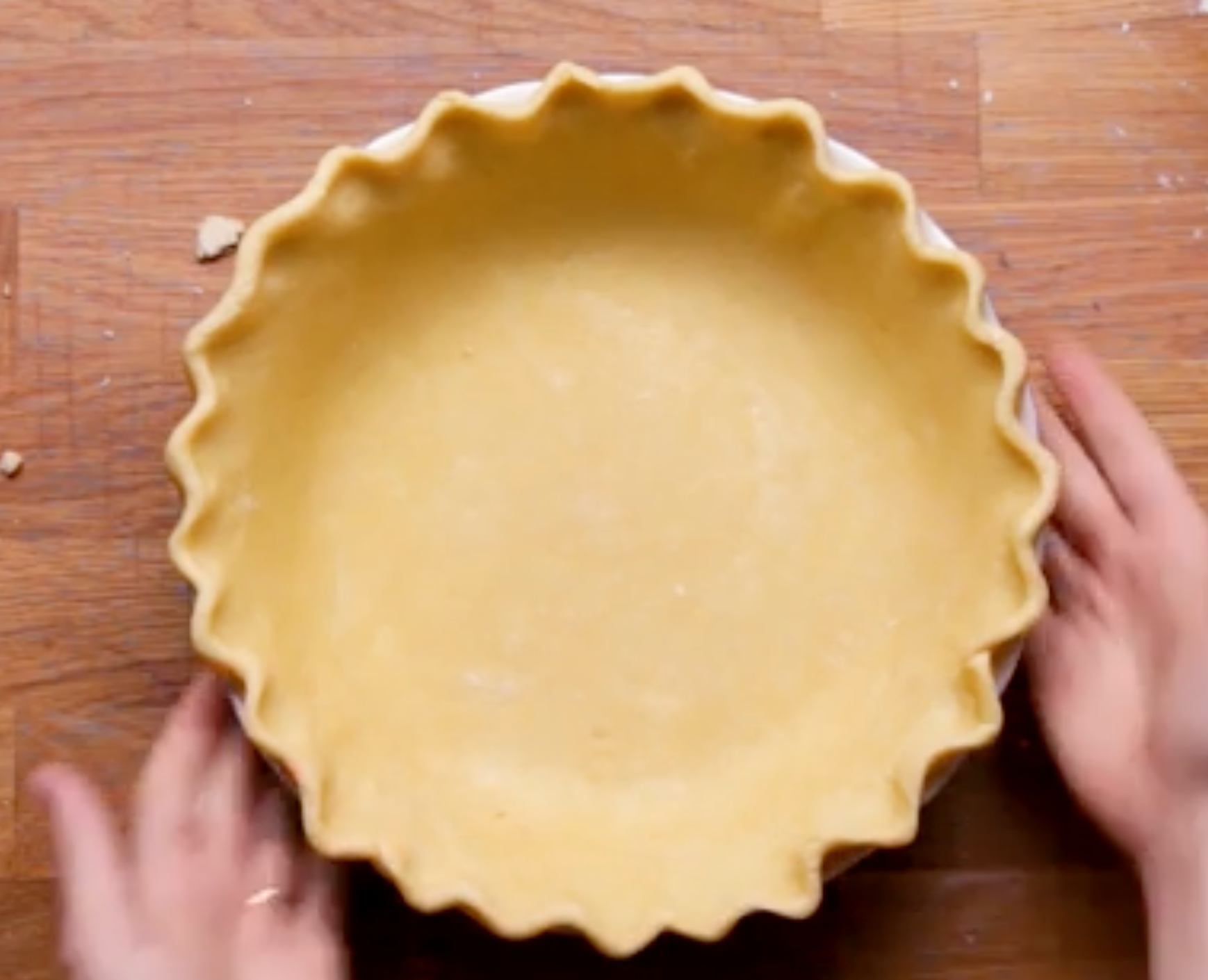 Hands shaping a pie crust with fluted edges in a dish on a wooden countertop