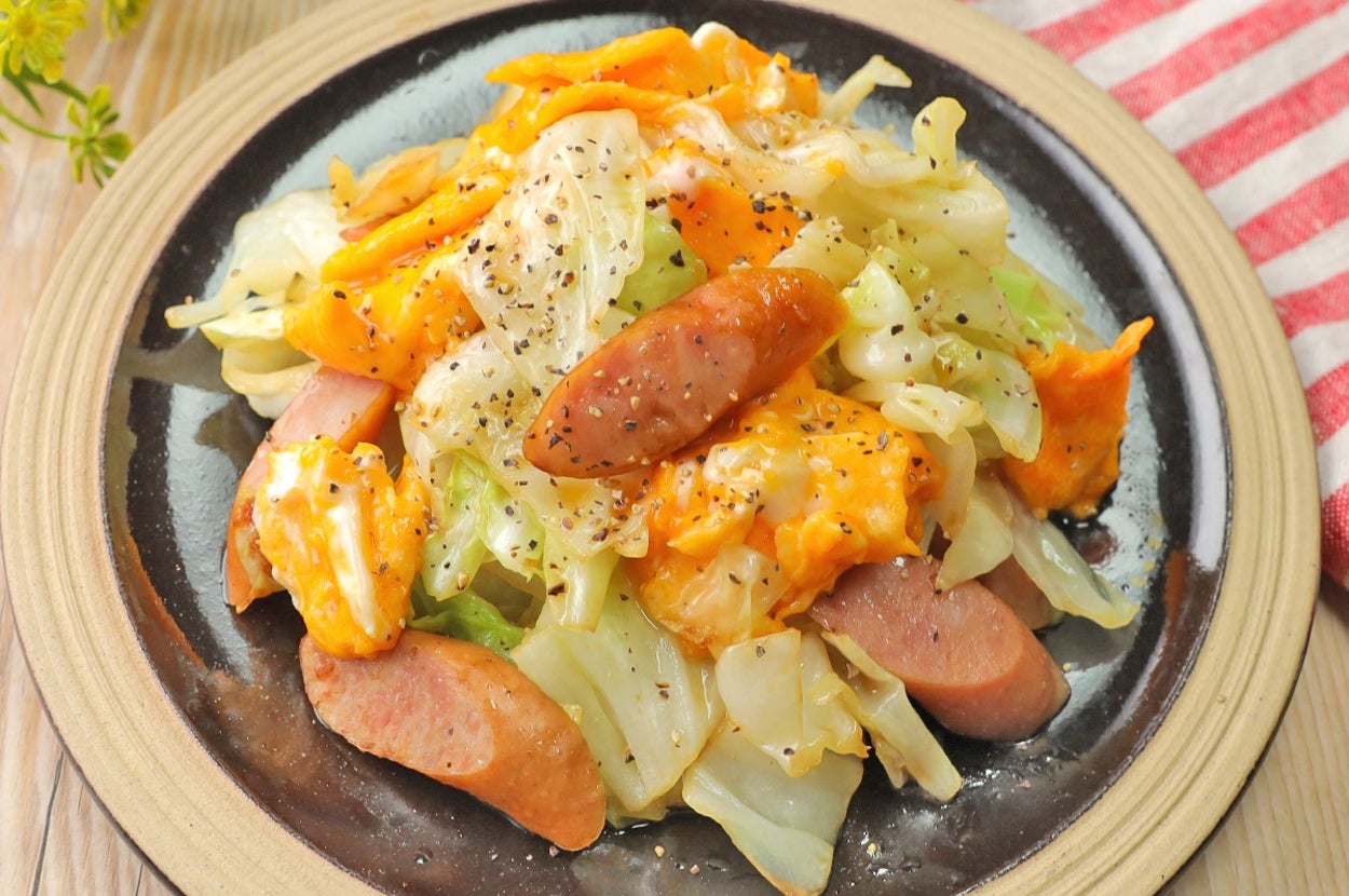 皿の上にキャベツとソーセージ、チーズの料理。上に黒胡椒がかかっている。木製のテーブルと赤白の布が背景。