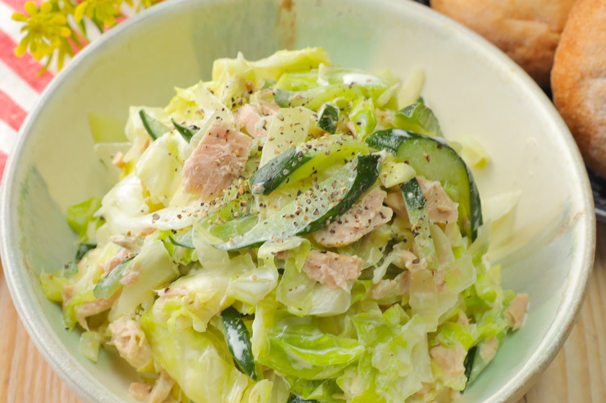 ボウルに盛られたキャベツ、きゅうり、ツナのサラダ。バックにパンが添えられたテーブルセッティング。