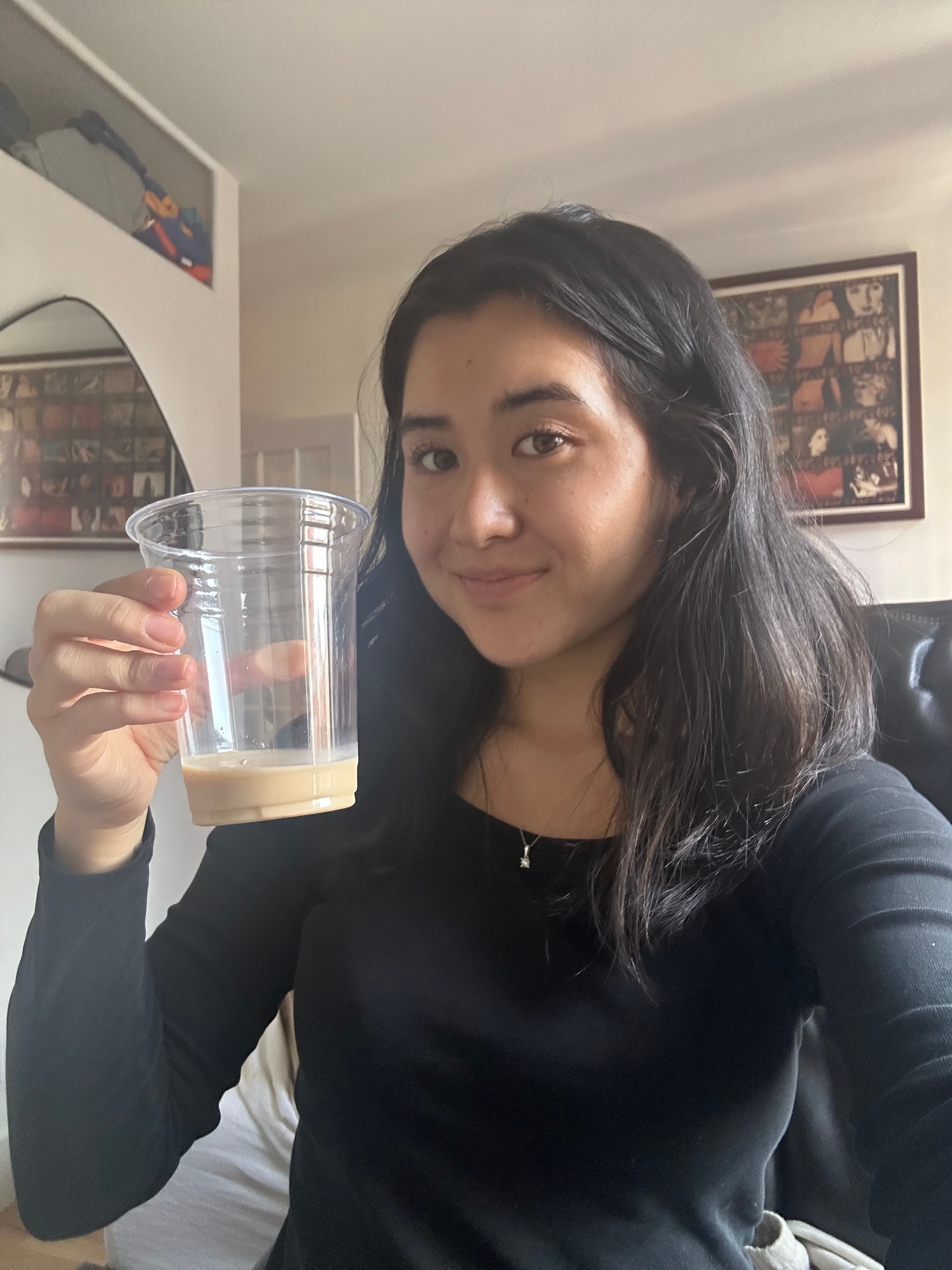 Person smiling indoors, holding a clear plastic cup with a small amount of beverage at the bottom
