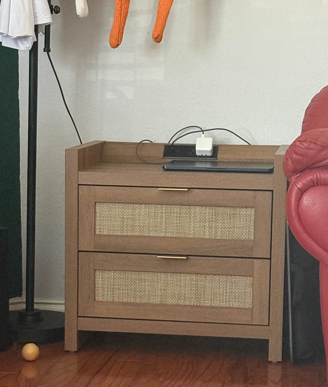 Wooden nightstand with 2  woven-front drawers, a telephone  and charger connected  top, adjacent  to portion  of a reddish  sofa  and a level  lamp