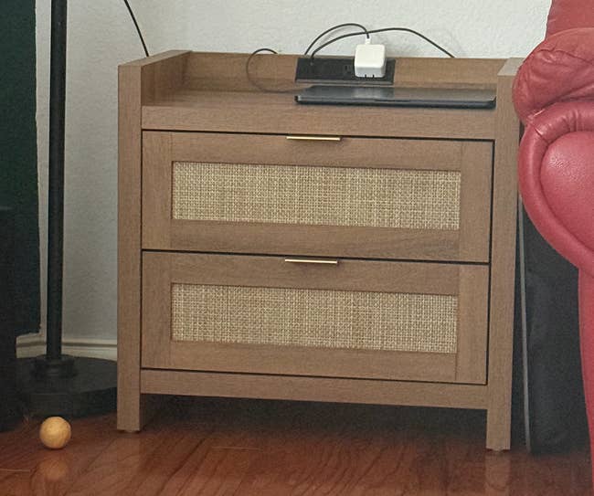Wooden nightstand with 2 woven-front drawers, a telephone and charger connected top, adjacent to portion of a reddish sofa and a level lamp