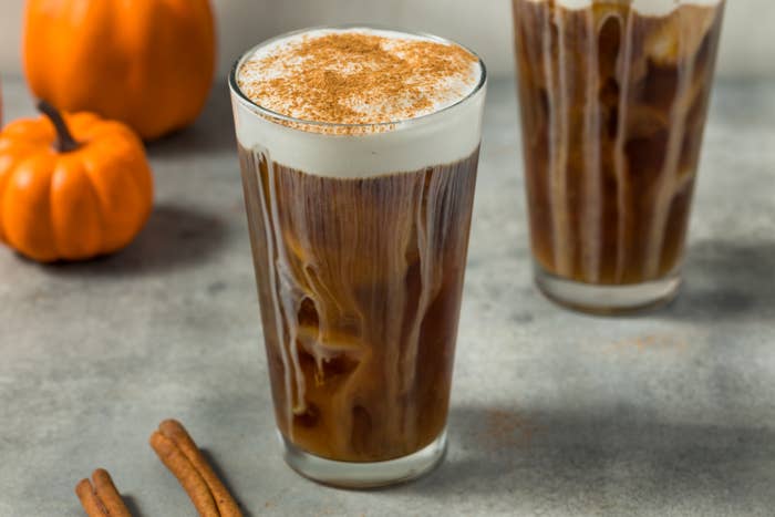 Two iced coffees with creamy foam on top, sprinkled with cinnamon. A small pumpkin and cinnamon sticks are nearby