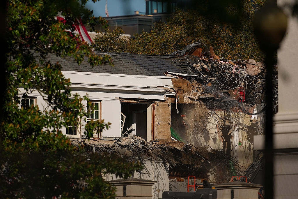 Partially demolished building with visible debris and exposed interior, surrounded by trees and construction equipment