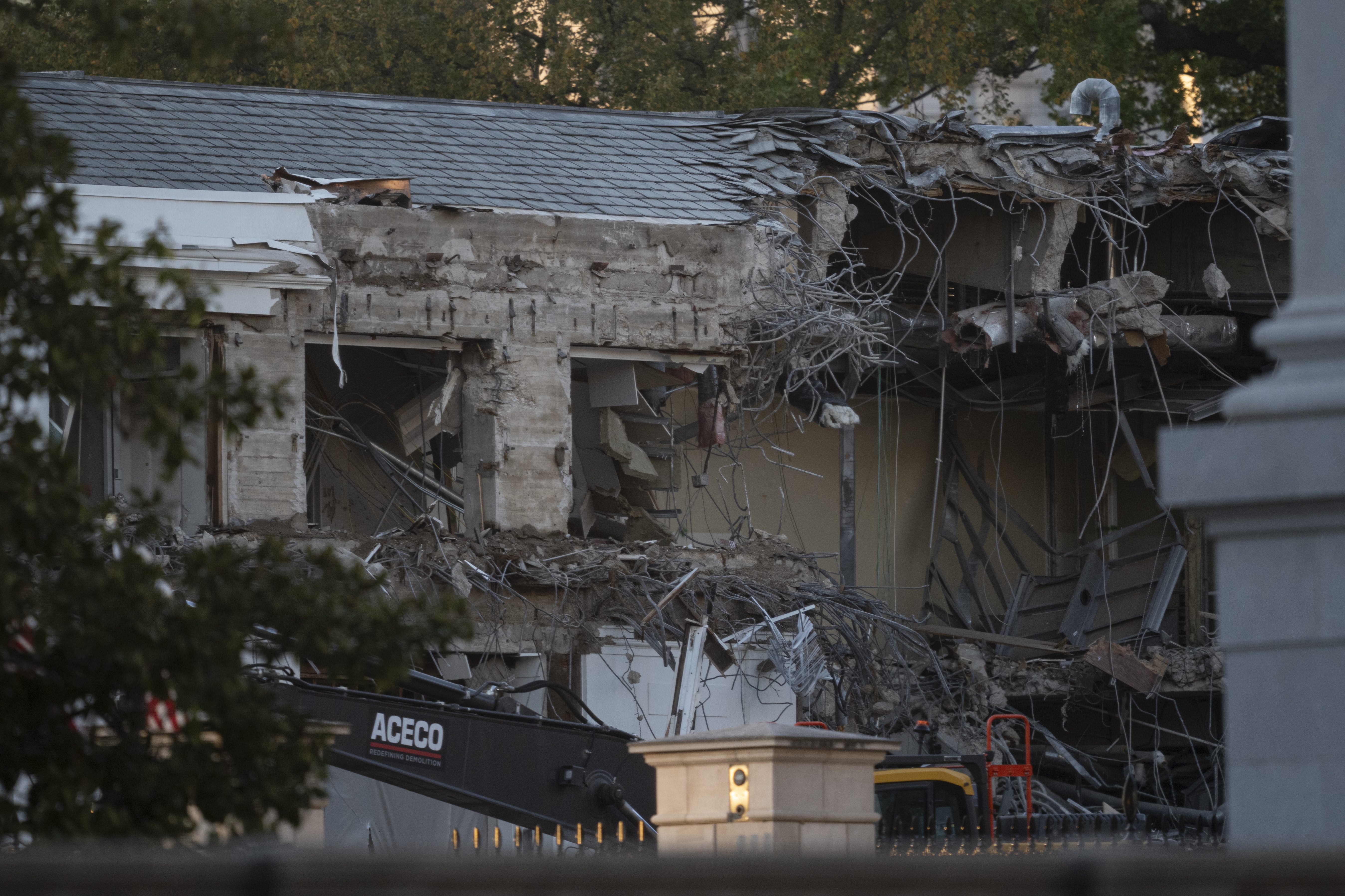Partially demolished building with exposed steel beams and debris, visible through trees and fencing