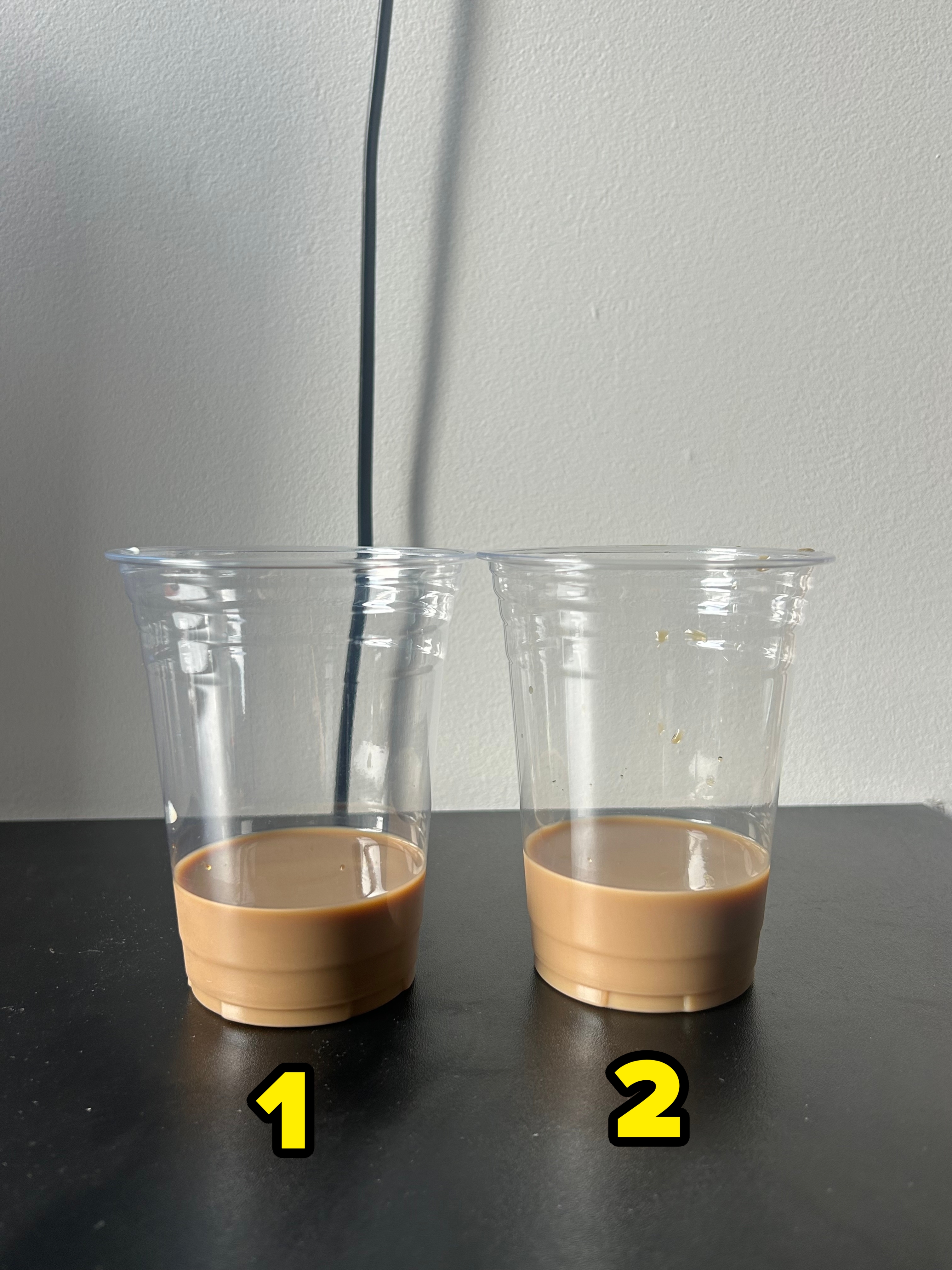 Two clear plastic cups on a black surface, each containing a small amount of coffee. A cable is visible against a plain wall in the background