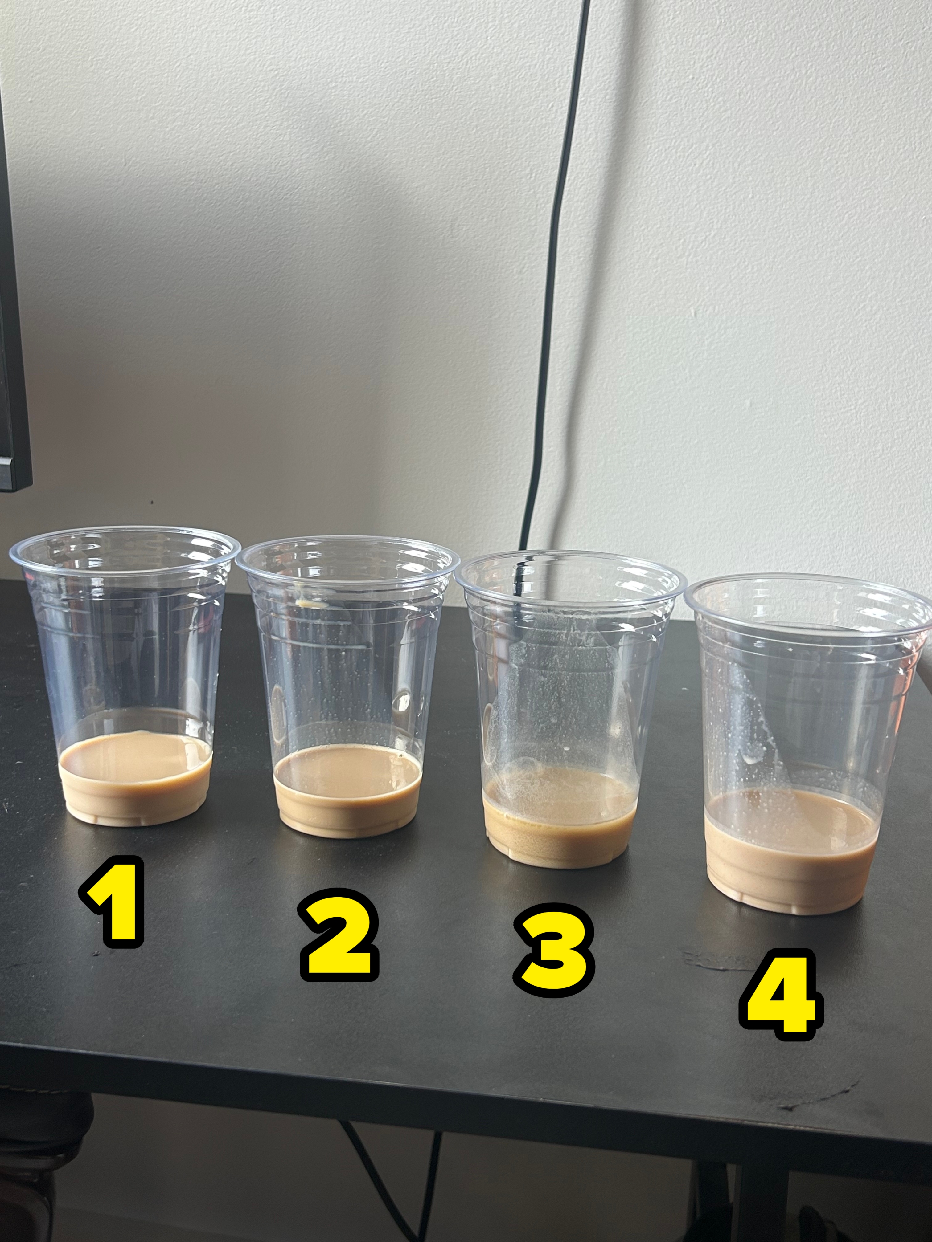 Four clear plastic cups with a small amount of beige liquid sit on a black table. Background shows a white wall with a black cable