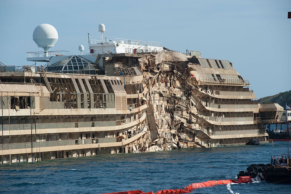 Damaged cruise ship partially submerged in water, showing extensive damage to its side. Emergency barriers float nearby