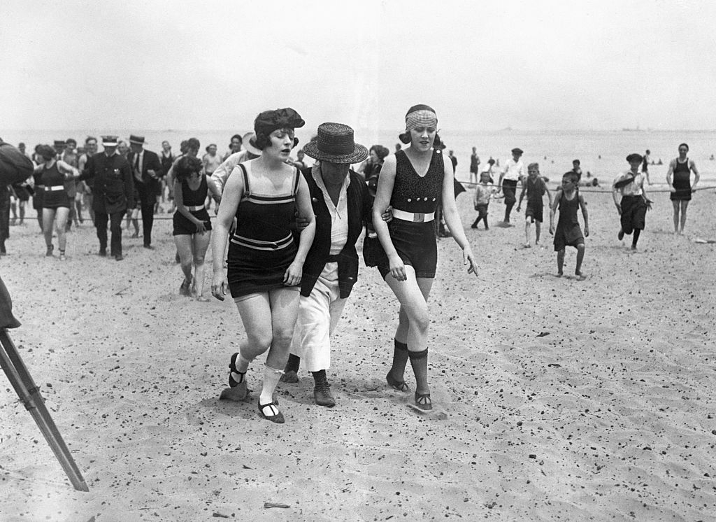 People in vintage swimwear on a crowded beach, one person helps another to walk