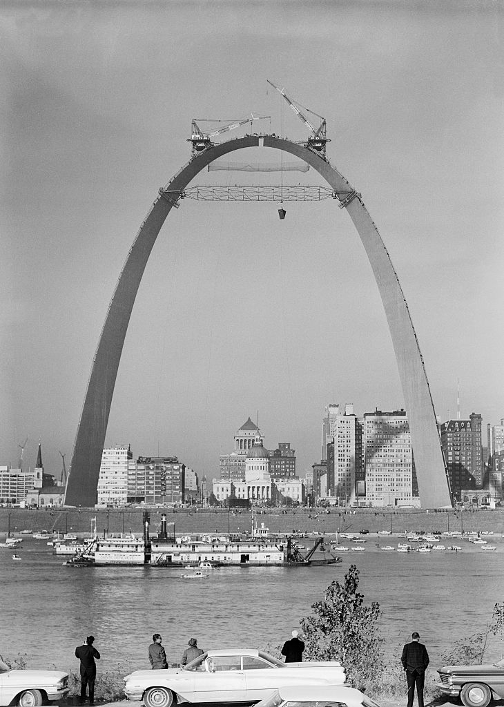 Construction of the Gateway Arch in progress, with cranes at the top and workers below, as people and cars gather to watch by the waterfront