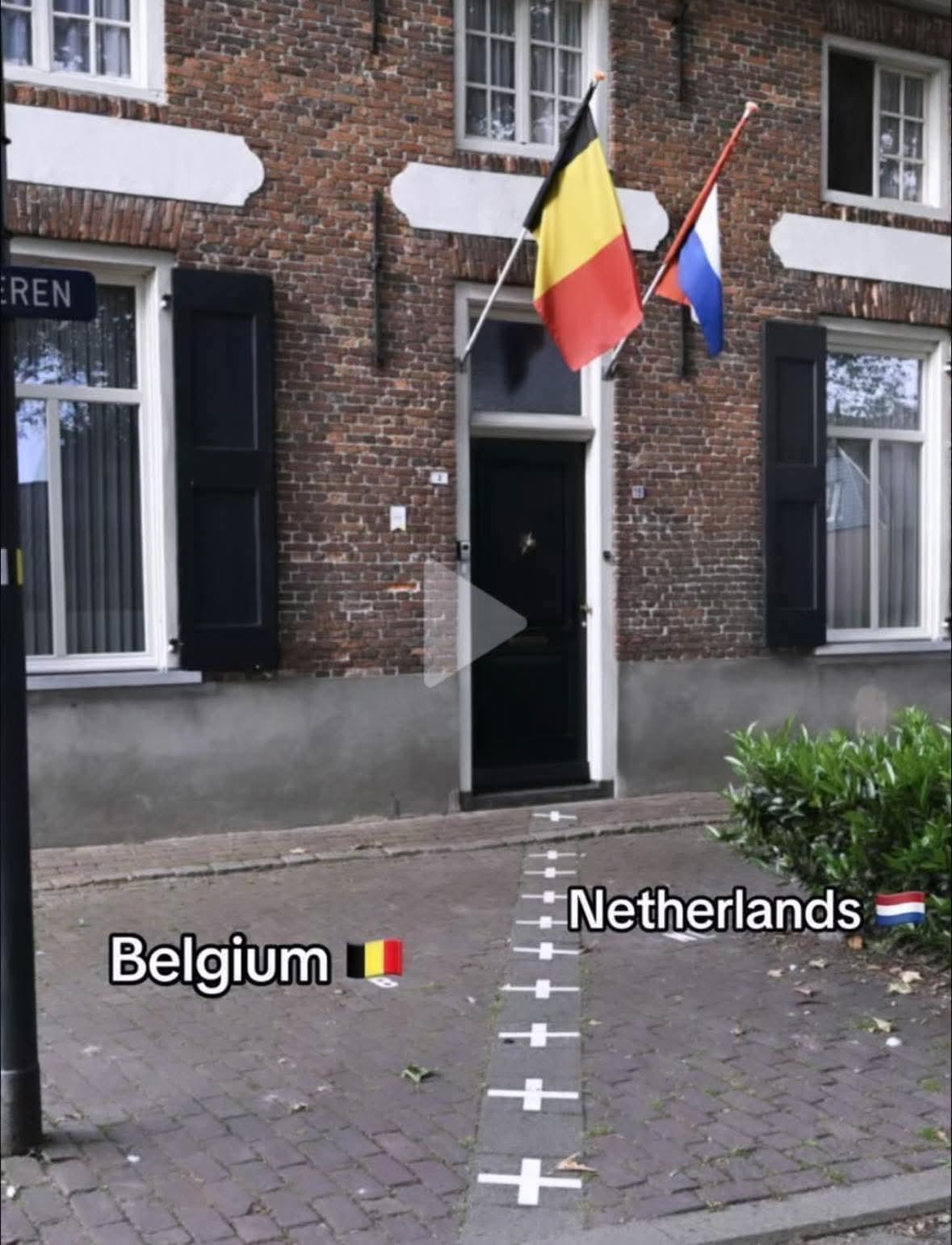 House with Belgian flag on the left and Dutch flag on the right. A border line runs across the sidewalk and doorstep, dividing Belgium and Netherlands