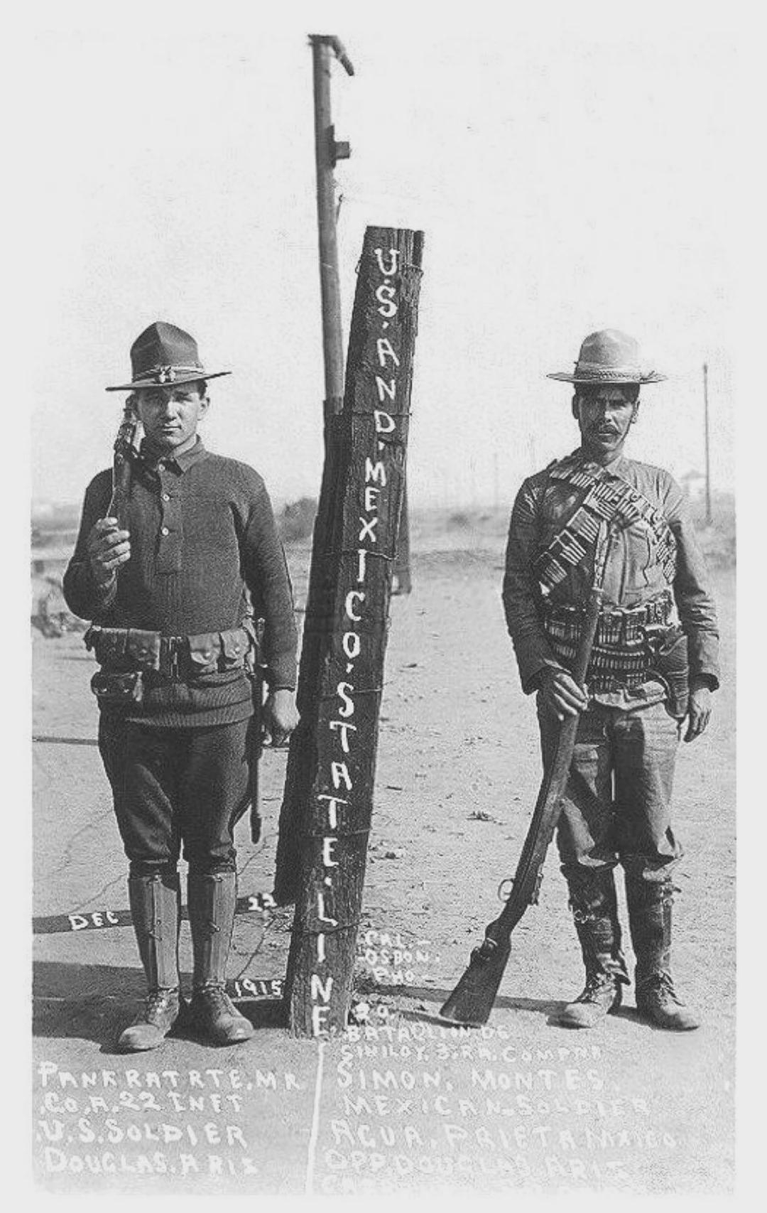 Two men in early 20th-century military uniforms stand on either side of a US and Mexico border marker, each holding rifles