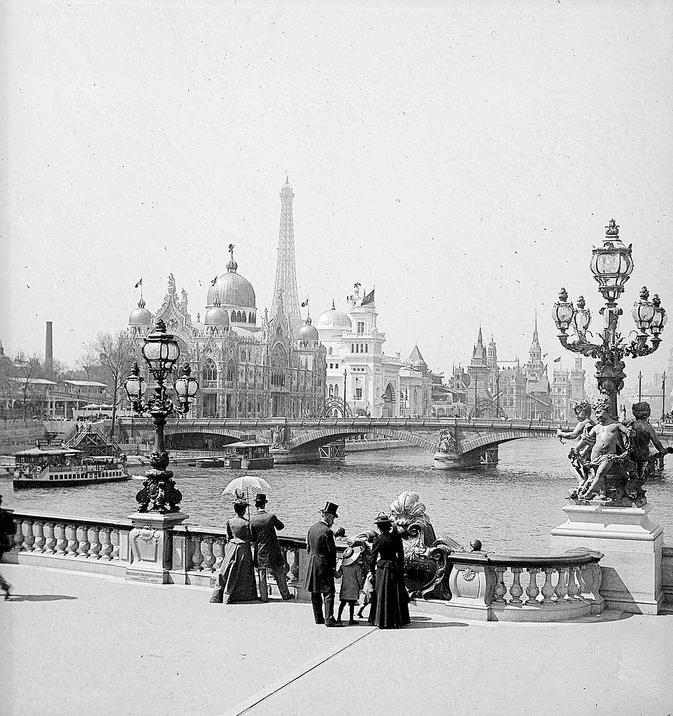 A historic scene of people in Victorian clothing overlooking a river with ornate bridges and distant exhibition towers