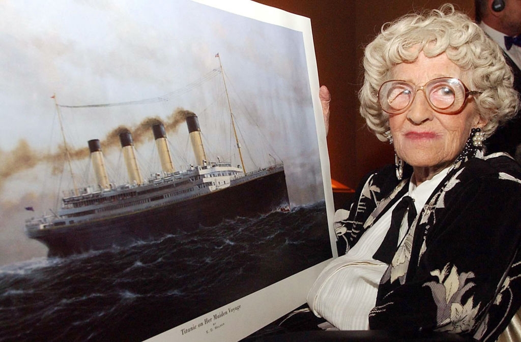 Elderly woman with glasses holding a large print of the Titanic; she's wearing a black and white patterned outfit