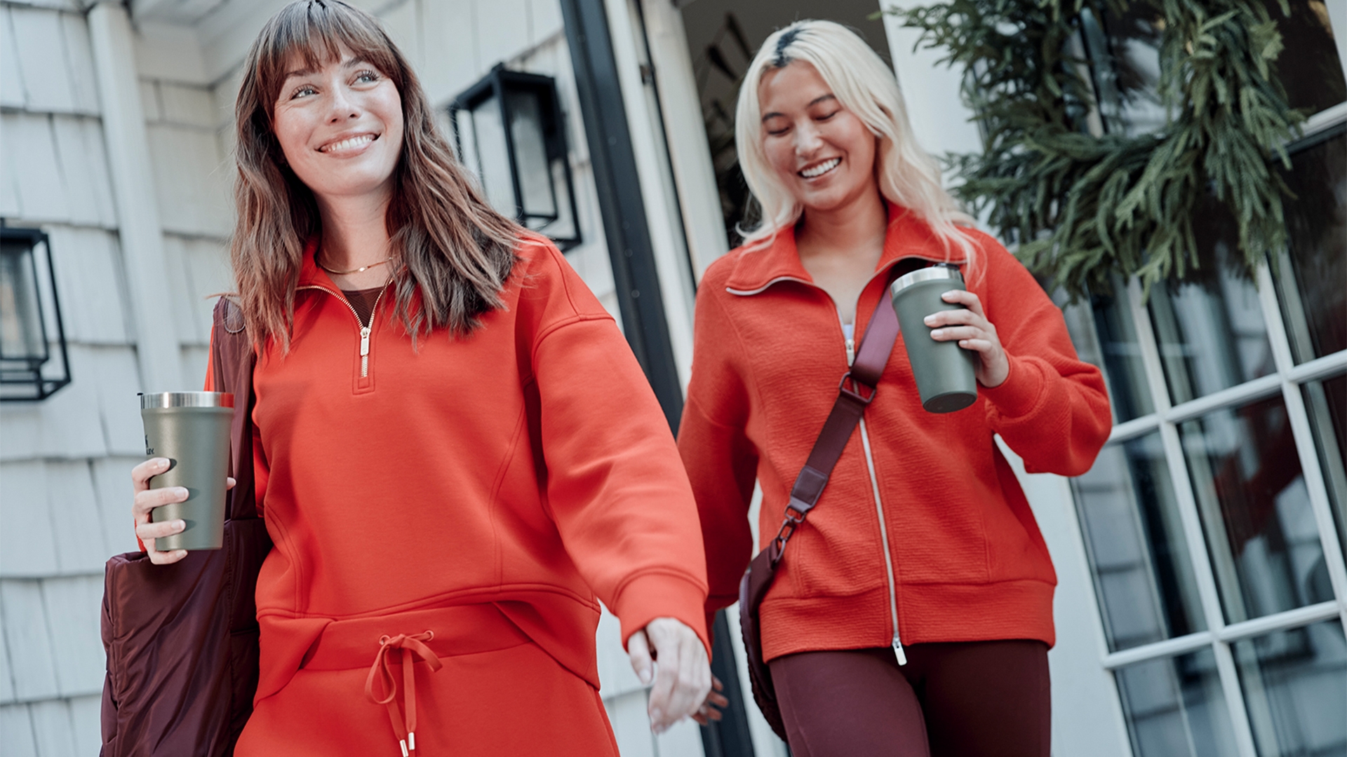 Two women smiling, walking outside holding beverages. They wear casual athleisure outfits with zippered tops, conveying a relaxed, active lifestyle