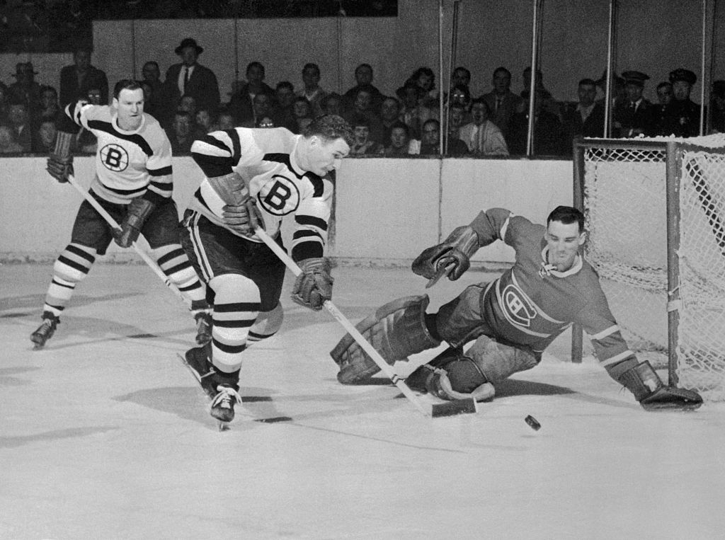Historic hockey game: a Boston Bruins player attempts to score against a Montreal Canadiens goalie who is defending the net. Spectators watch intently
