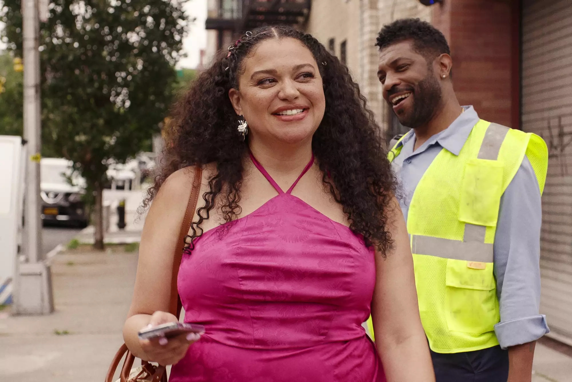 Michelle Beautau smiling successful a sleeveless formal holding a telephone passes a postulation defender connected the sidewalk successful "Survival of the Thickest"