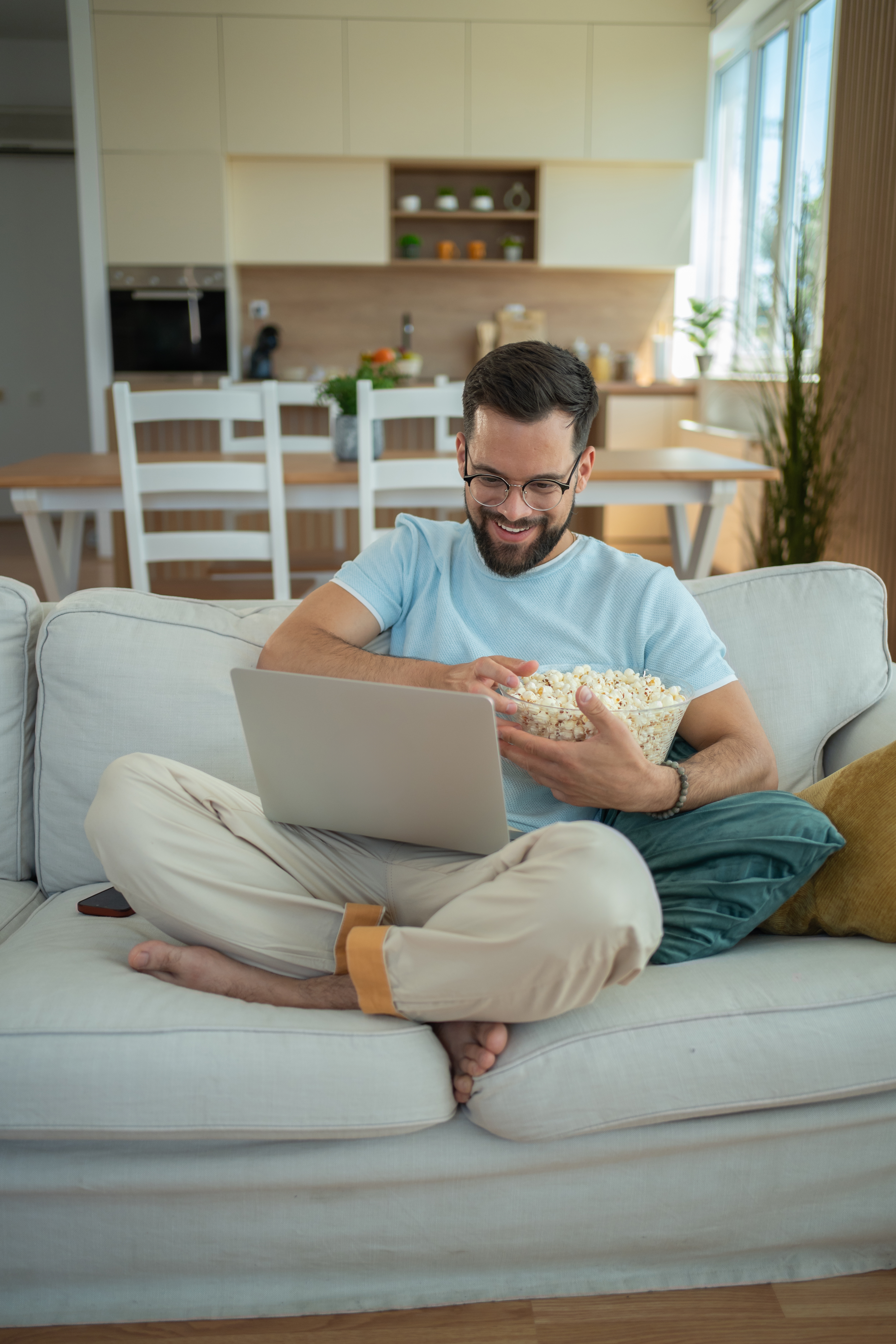 Mann som sitter på sofaen med laptop, smiler og spiser popcorn i en moderne stue