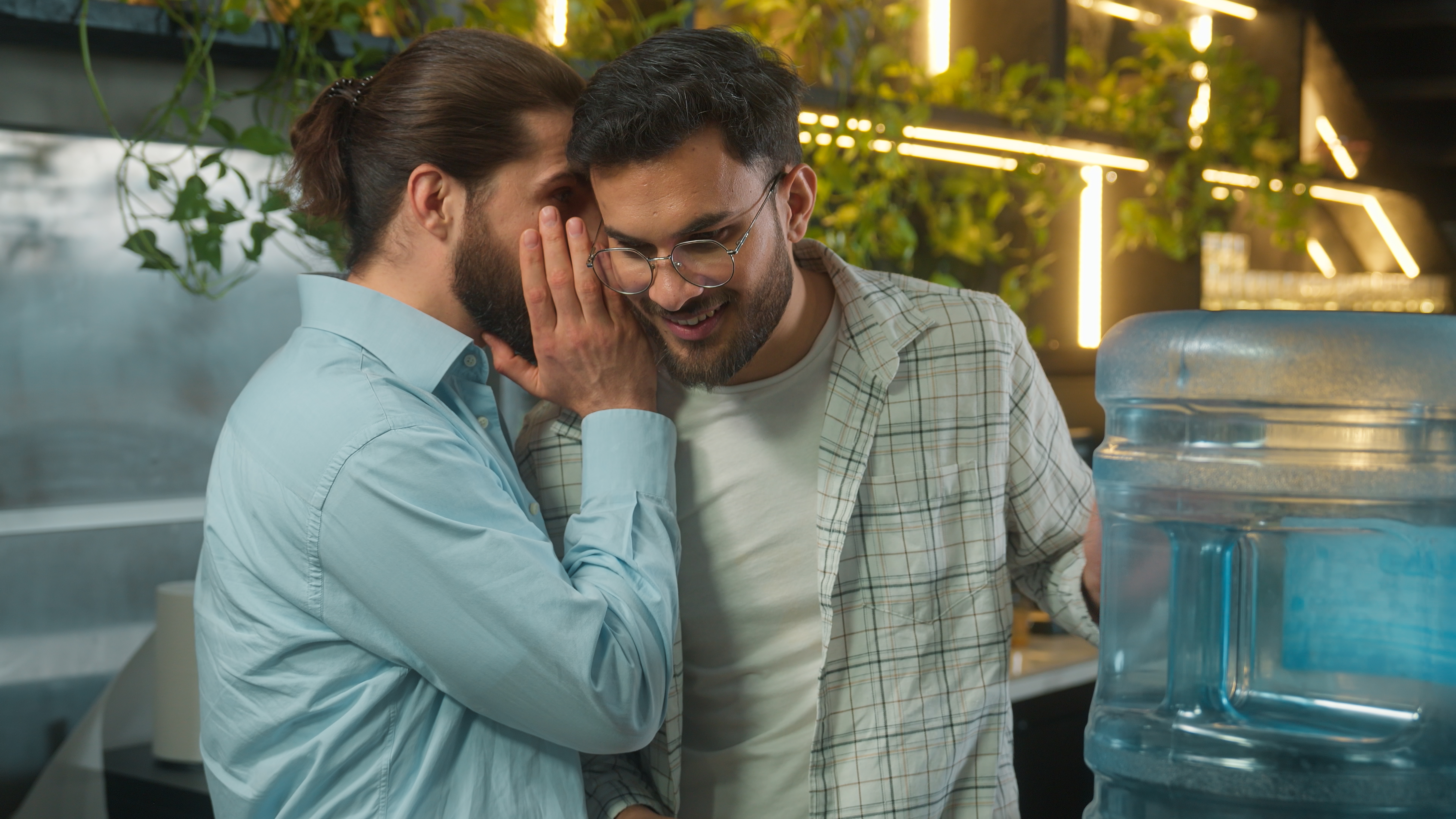 Two men astatine  a h2o  cooler. One whispers to the other, who smiles. Indoor mounting  with plants and a modern   background