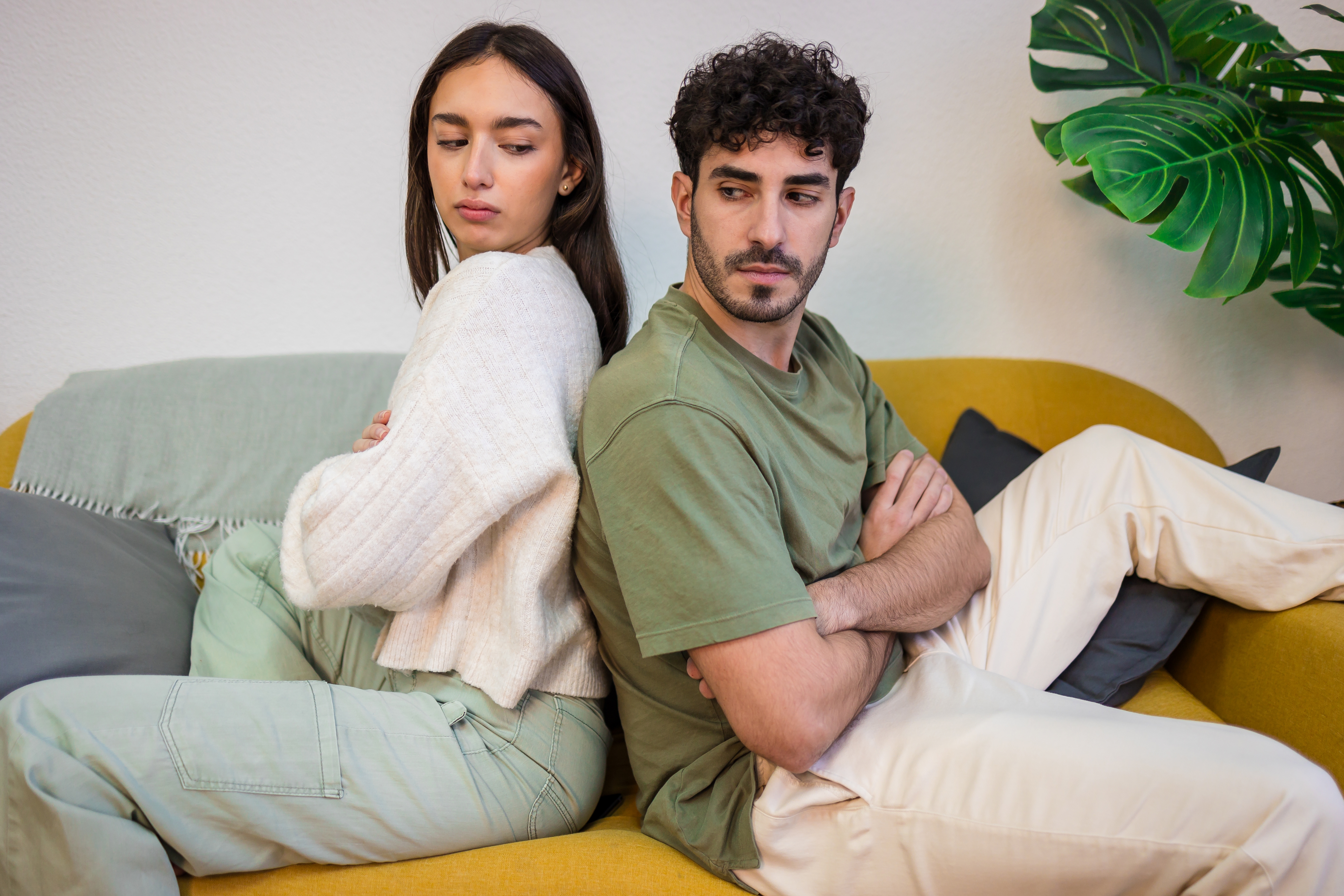 Two individuals beryllium   back-to-back connected  a sofa, arms crossed, some  with pensive expressions