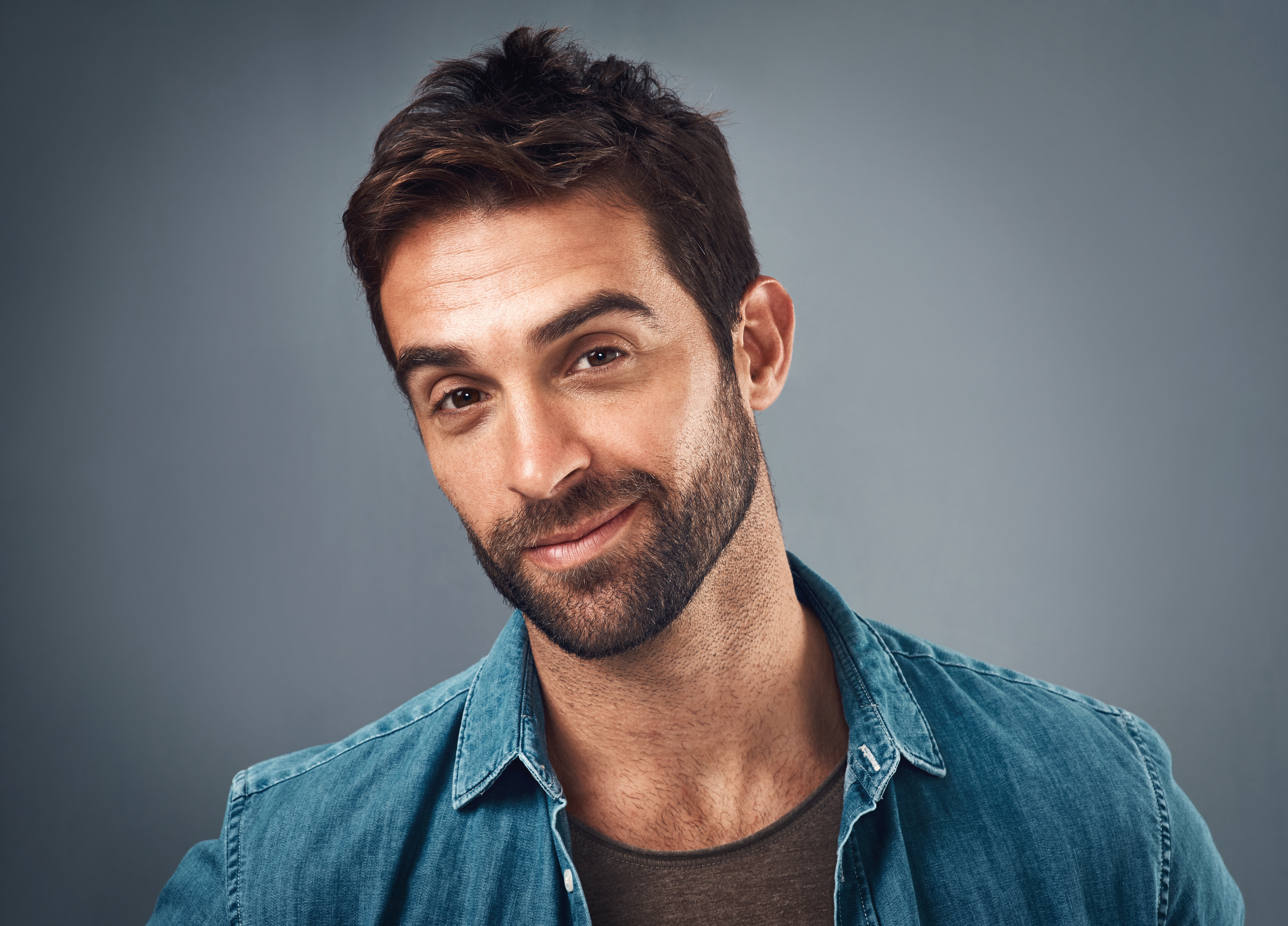 Man with abbreviated  hairsbreadth  and a beard, smiling slightly, wearing a denim garment  implicit    a t-shirt against a plain background