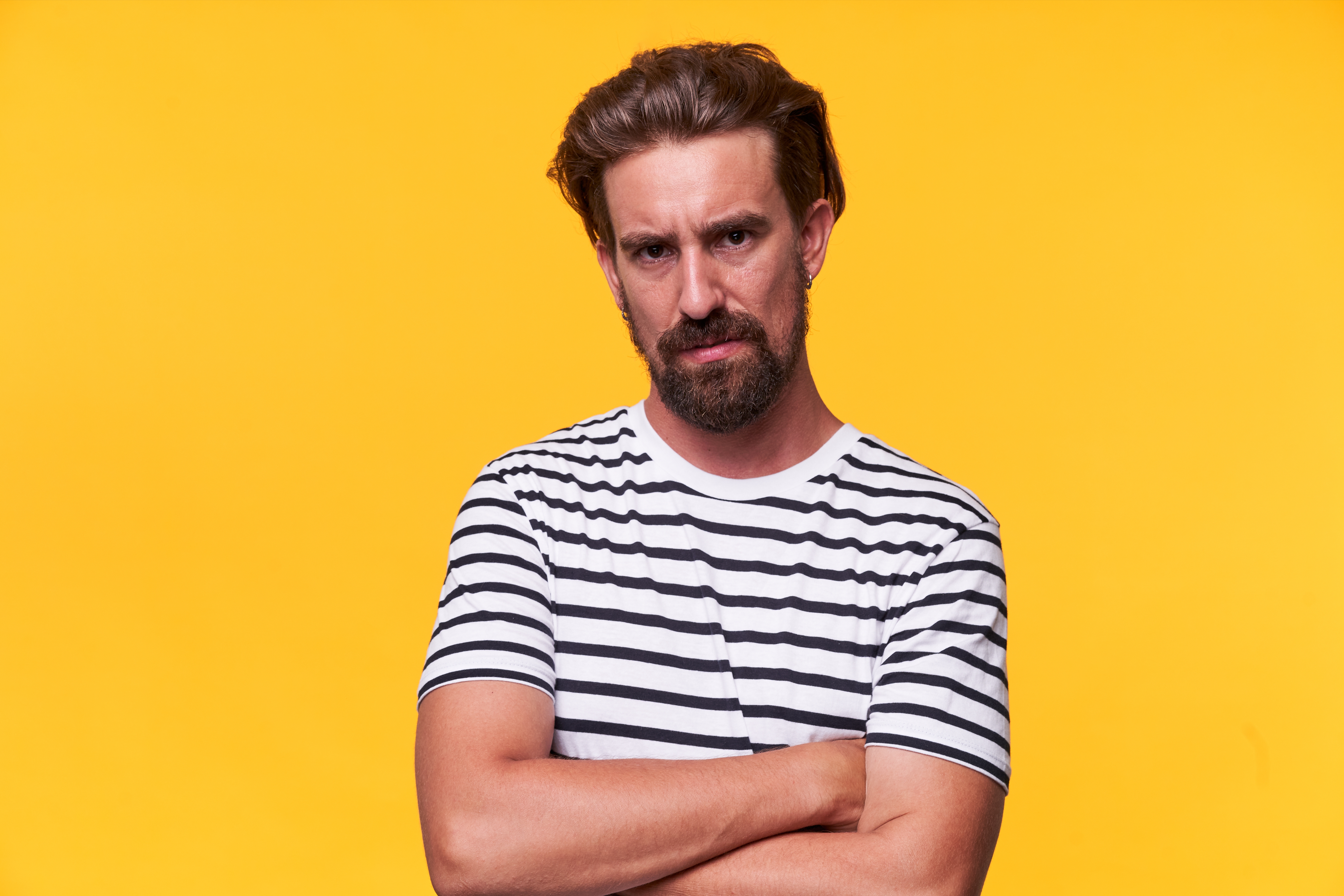 A idiosyncratic   with a beard looks serious, arms crossed, wearing a striped garment  against a plain background