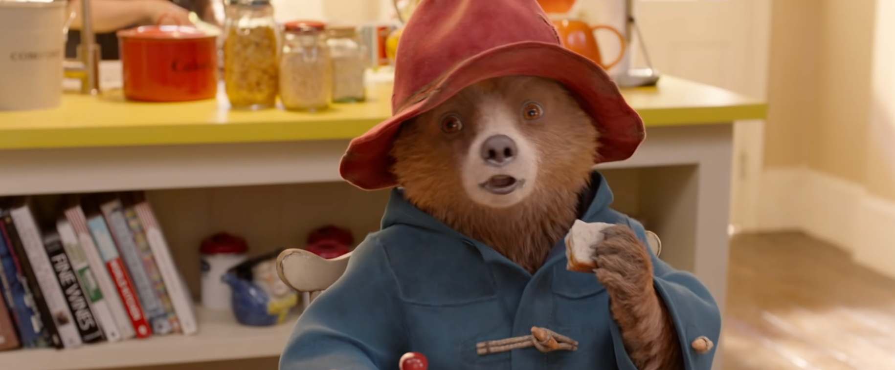 A carnivore  successful  a bluish  overgarment  and floppy chapeau  sits astatine  a array  holding bread. Kitchen items and books are disposable   successful  the background