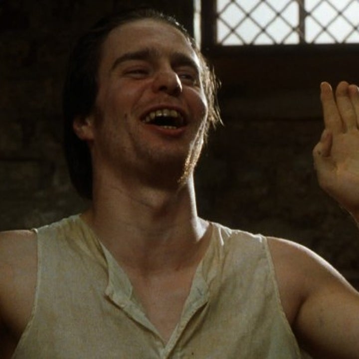 Man in a rustic setting, wearing a sleeveless, loose top, raises both hands while smiling, suggesting joy or surprise