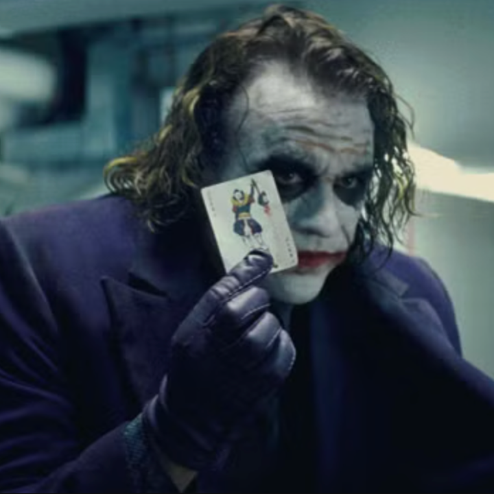 Character in dark suit holding a Joker card up to face, inside an industrial setting, with an intense expression