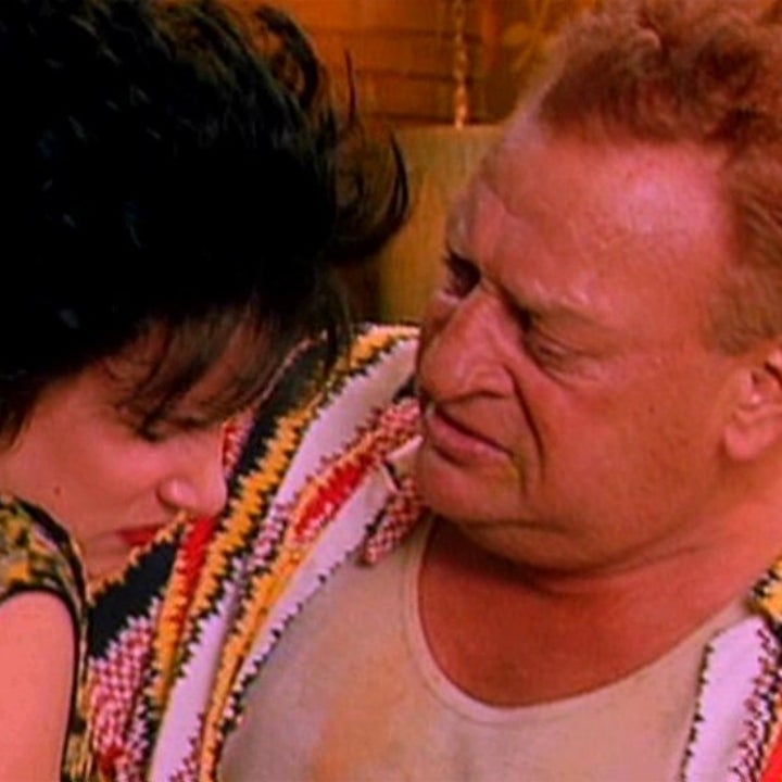 A man wearing a tank top and knit cardigan looks concerned while speaking to a woman with short hair wearing a patterned top
