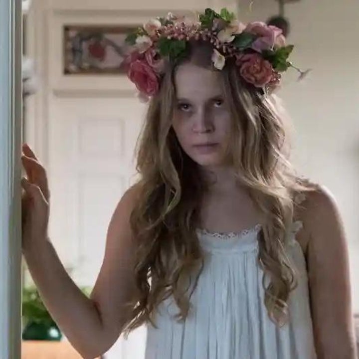 Person with long hair wearing a floral crown and white dress stands indoors, looking forward with a serious expression