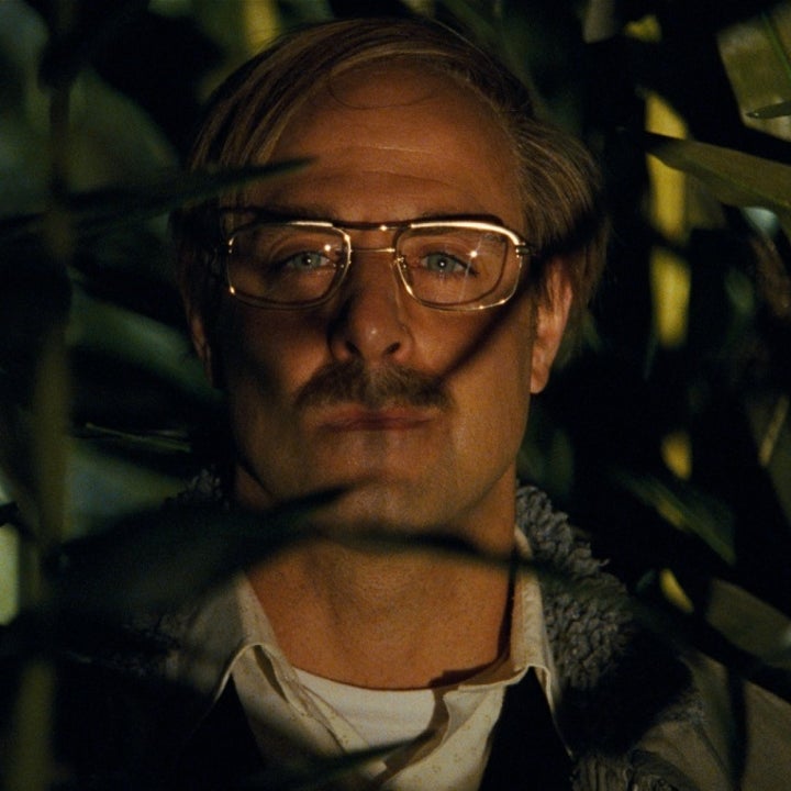 A man with glasses and a mustache is partially obscured by leaves, creating a mysterious, contemplative look