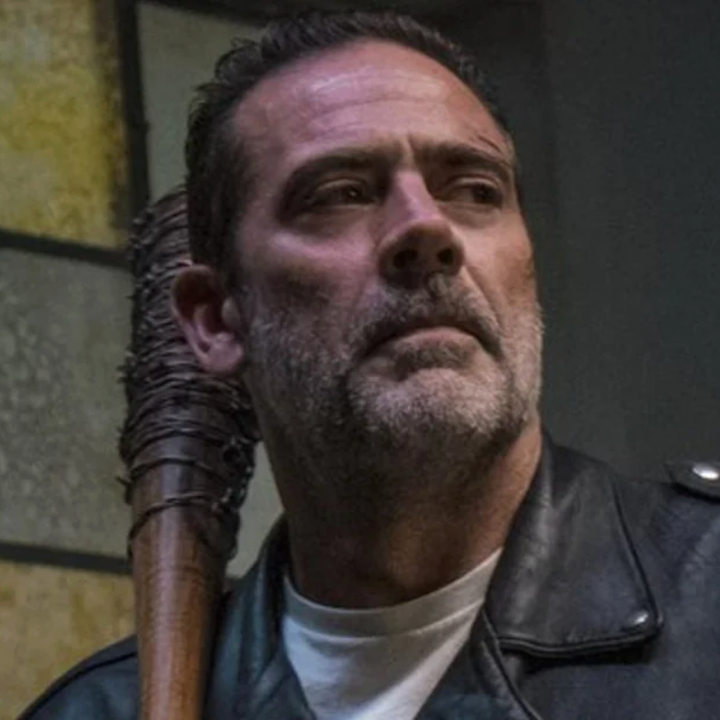 A person holding a barbed wire-covered bat, wearing a leather jacket and looking off to the side in a dramatic scene