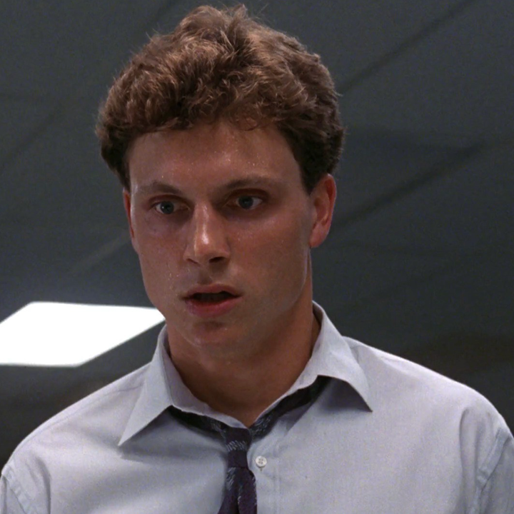 A surprised man stands indoors, wearing a dress shirt and a loosened tie
