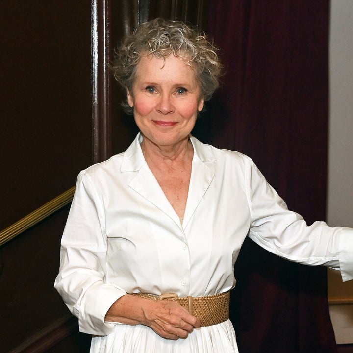 Person in a classic white dress with a pleated skirt and tan woven belt, smiling at an indoor event