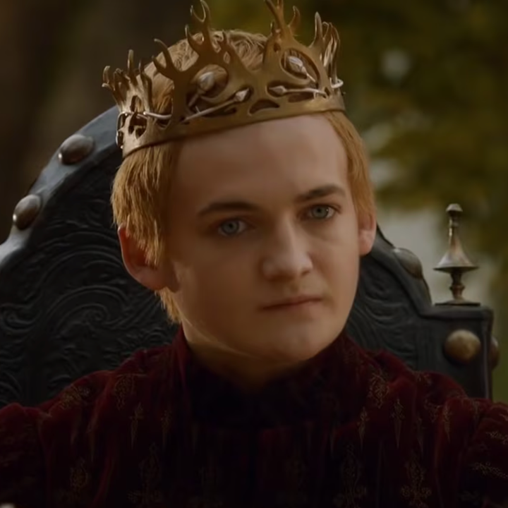A person wearing a gold crown sits on a throne, looking forward with a serious expression. They are dressed in ornate medieval-style attire